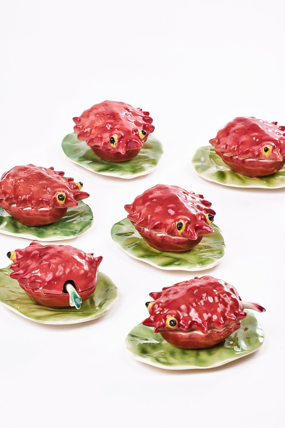 Six glossy red ceramic fish-shaped cocottes with yellow-and-black eyes, each resting on a green leaf-shaped plate against a white background.