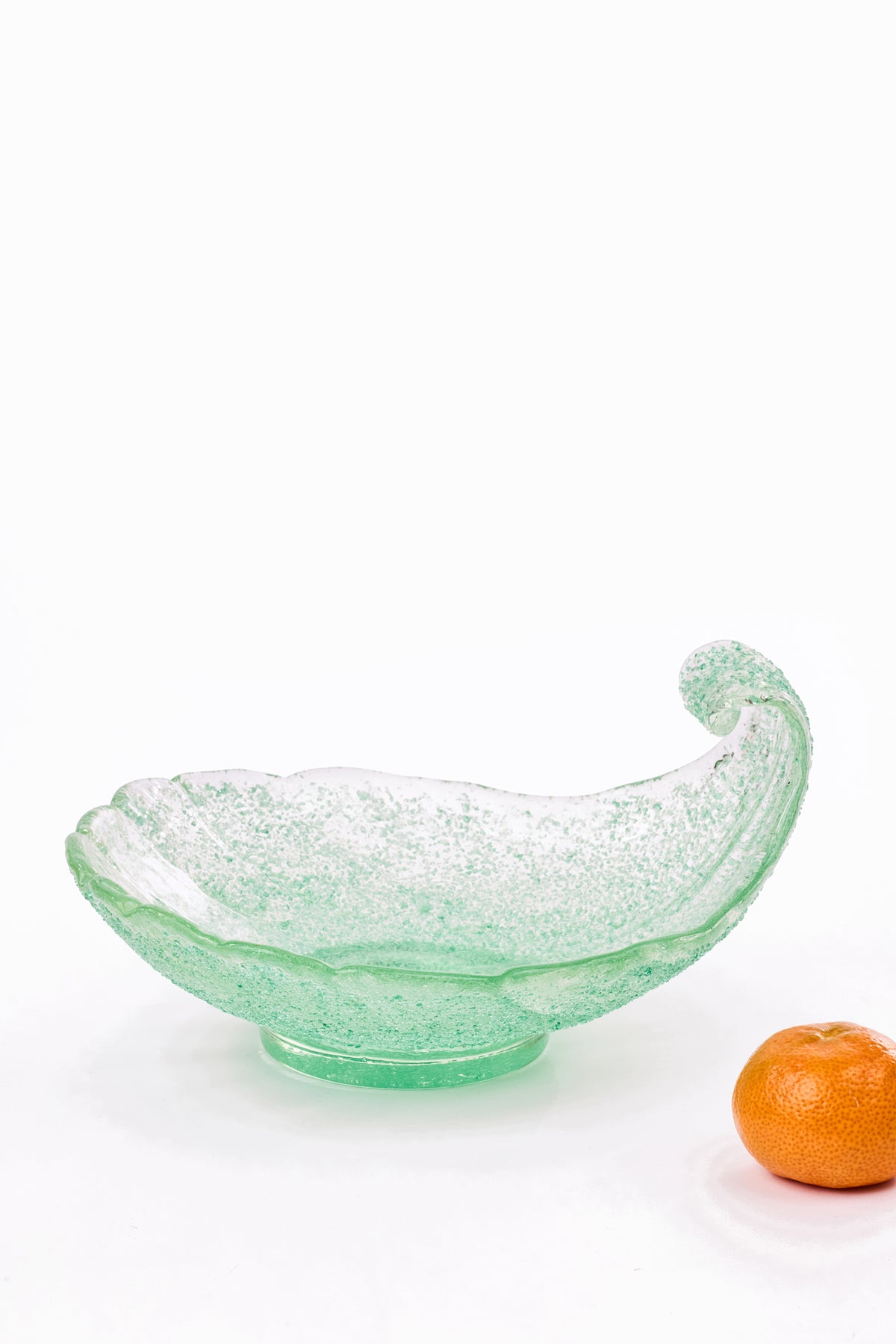 A light green glass bowl with a textured, speckled finish and a curved, shell-like rim, shown on a white background next to a small orange fruit.