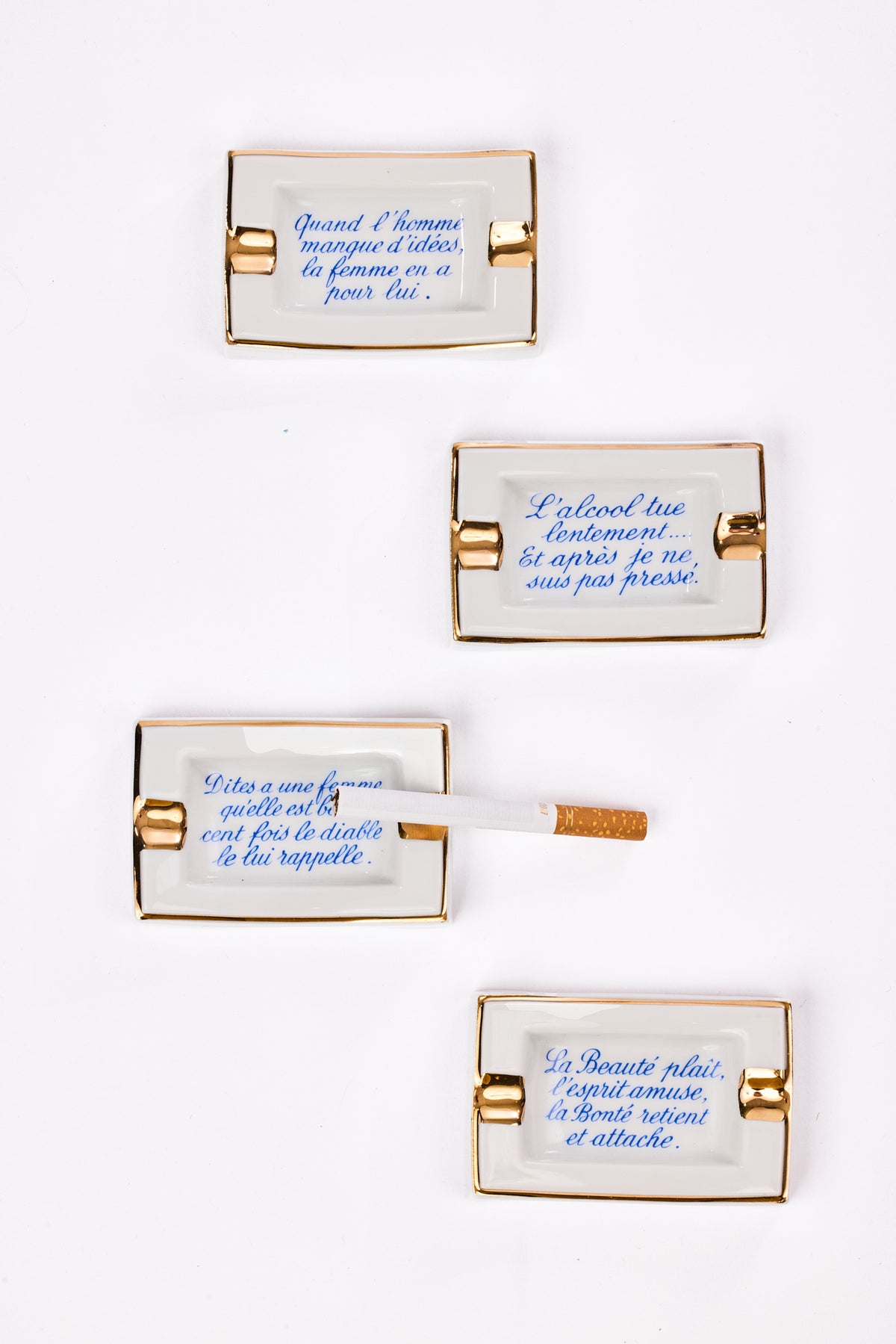 Four rectangular white porcelain cigarette cases with gold metal clasps and blue French text, arranged vertically on a white background. One case is shown open with a cigarette protruding.