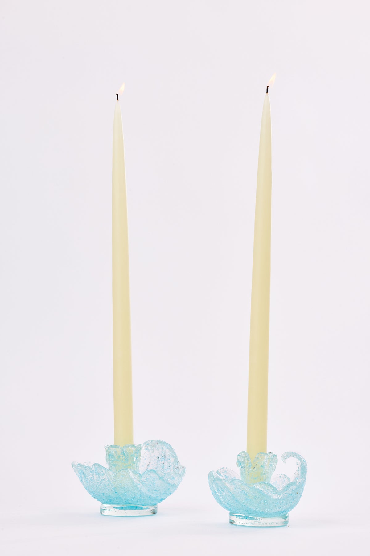 Two cream taper candles standing in light blue textured glass holders in the shape of a shell against a plain white background.