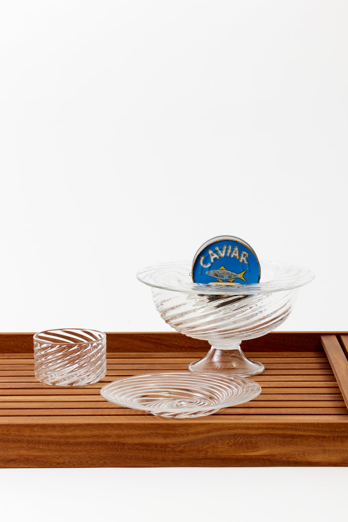 Party-sized clear artisanal twisted handblown glass caviar service, blini plate, and creme fraiche dish resting on a wooden serving tray. Inside the service is a can of caviar. 