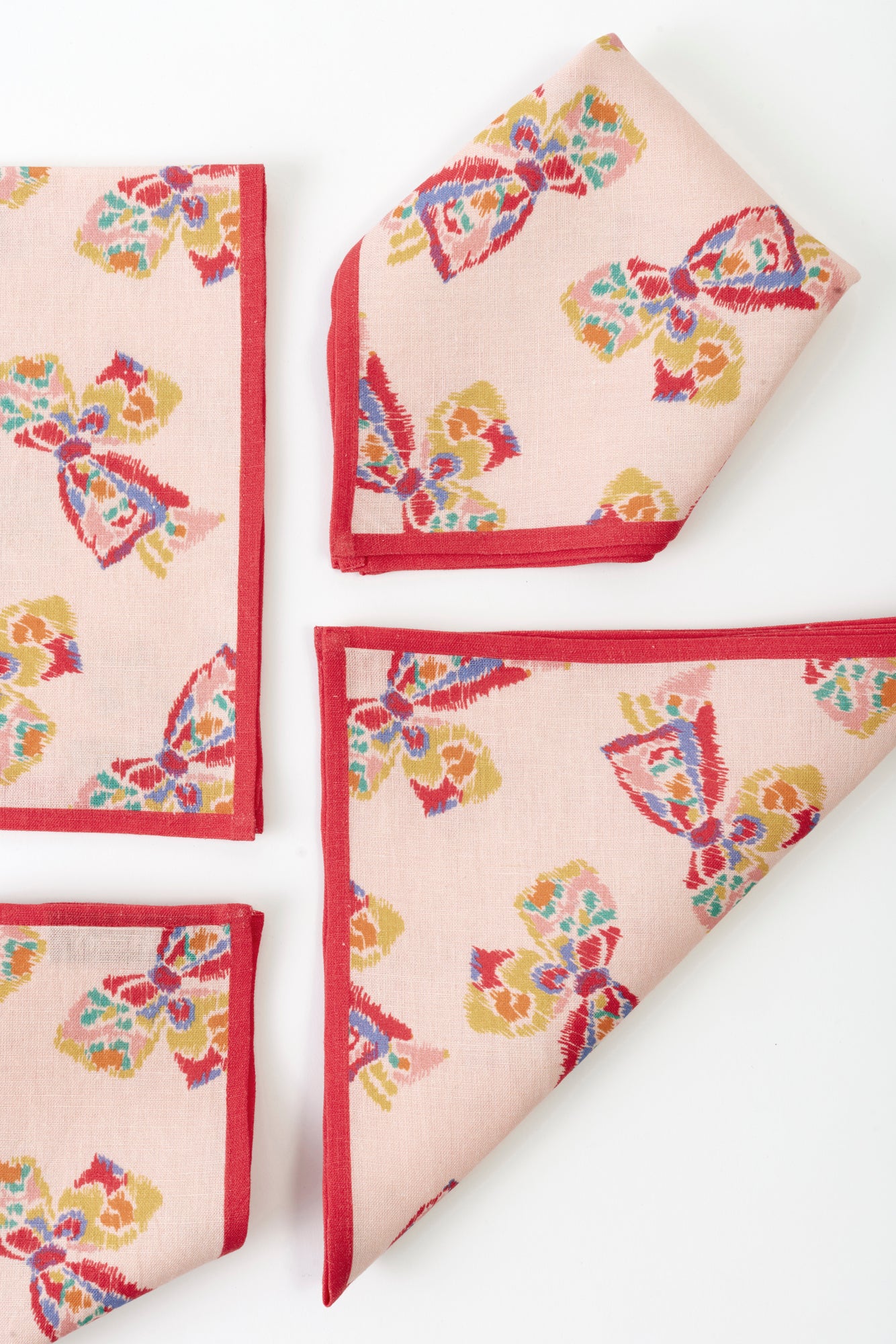 Four napkins placed on a white background. Each napkin is pink with a red border and features a printed pattern of a vintage-inspired rainbow bow. 