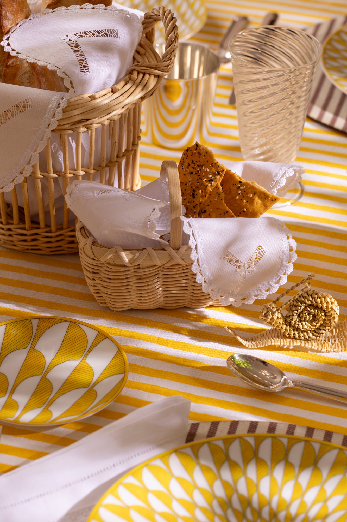 Close up photo of a table setting with a yellow and white striped tablecloth, yellow dishes, wicker breadbaskets with openwork embroidered linen fabrics, hand blown glass goblets, sterling silverware, and a corn husk snail.