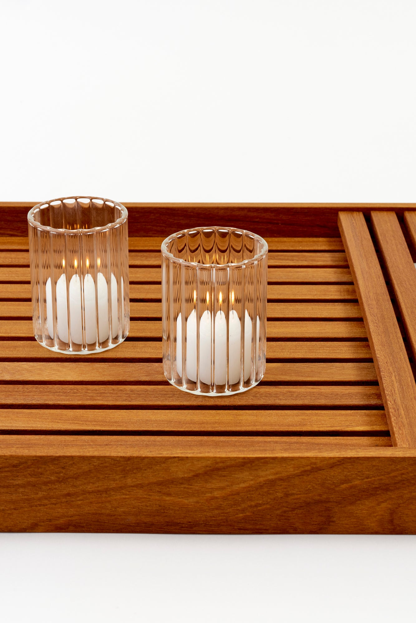 Two votive glasses on slatted wooden tray. Votives are clear fluted glass and have lit tea candles inside. Votives are 2.25" in diameter by 3.25" tall. 