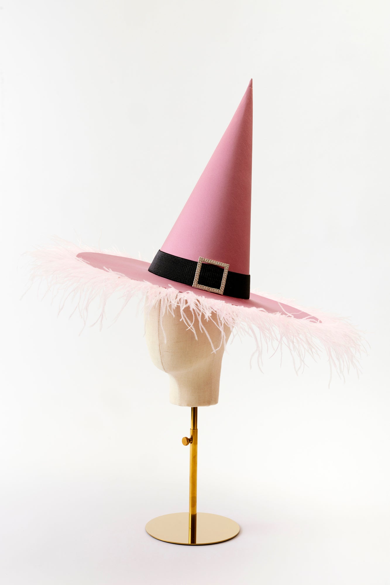 A glamorous pink witch hat with a tall, pointed top and wide brim adorned with delicate feather trim. The hat features a black band with a rhinestone buckle, adding a chic and whimsical touch to this fashionable accessory.