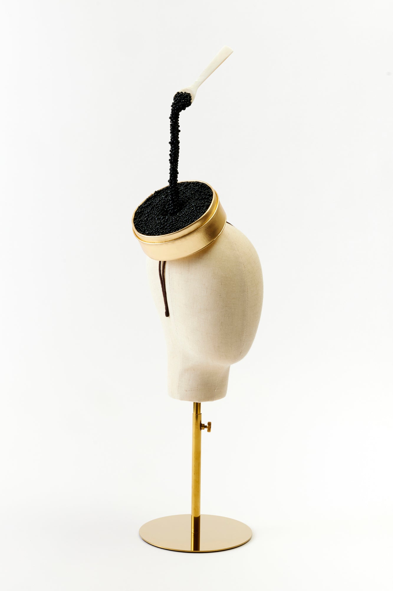 A quirky and luxurious hat resembling an open tin of black caviar. The gold-edged base holds black beads spilling over the side, while a tiny white spoon hovers over the caviar, creating a surreal and high-fashion accessory.