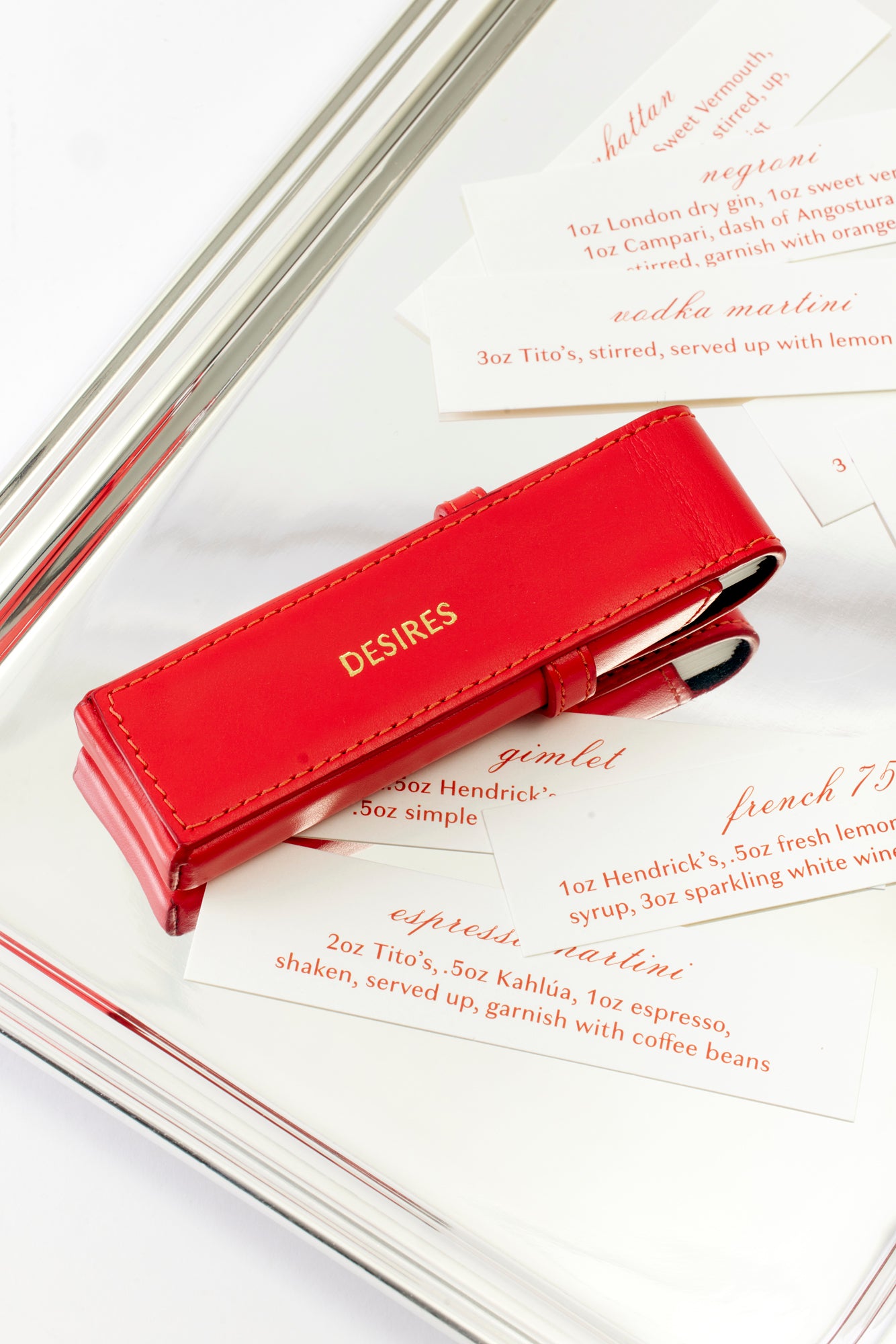 A luxurious red leather case embossed with the word 'DESIRES' rests on a polished silver tray surrounded by elegant cocktail recipe cards. Featuring classic drinks like the Martini and Negroni, this chic bar setup exudes sophistication and indulgence, perfect for upscale entertaining.