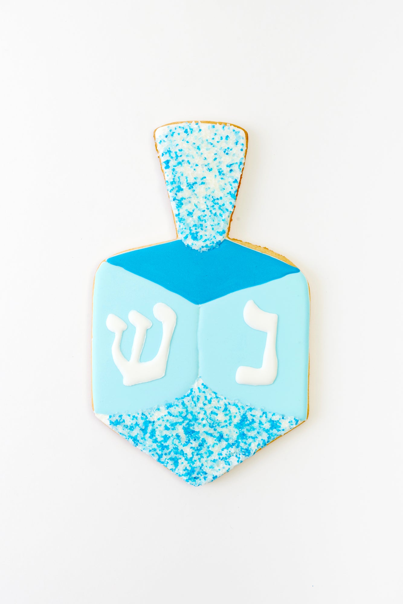 A dreidel-shaped cookie decorated in blue and white icing with sugar crystals at the top. The sides feature Hebrew letters in white, bringing a festive touch to this Hanukkah-inspired treat.