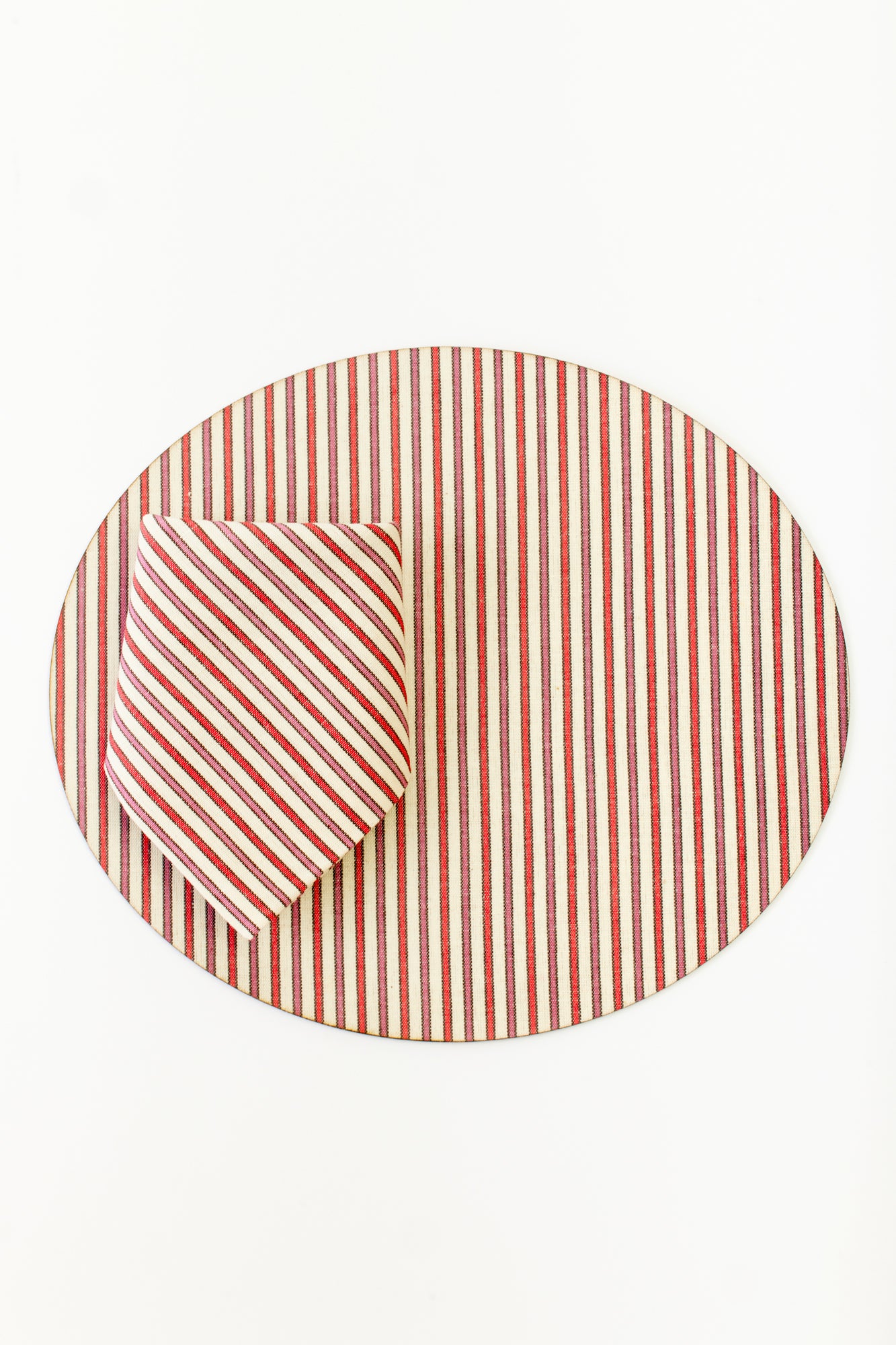 Old fashioned hardback placemat topped with a heavy duty blend of 60% linen, 40% cotton that has been coated for indestructibility. Oval shaped placemat with red, pink, and white vertical stripes. Paired with napkin with red, pink, and white stripes.  