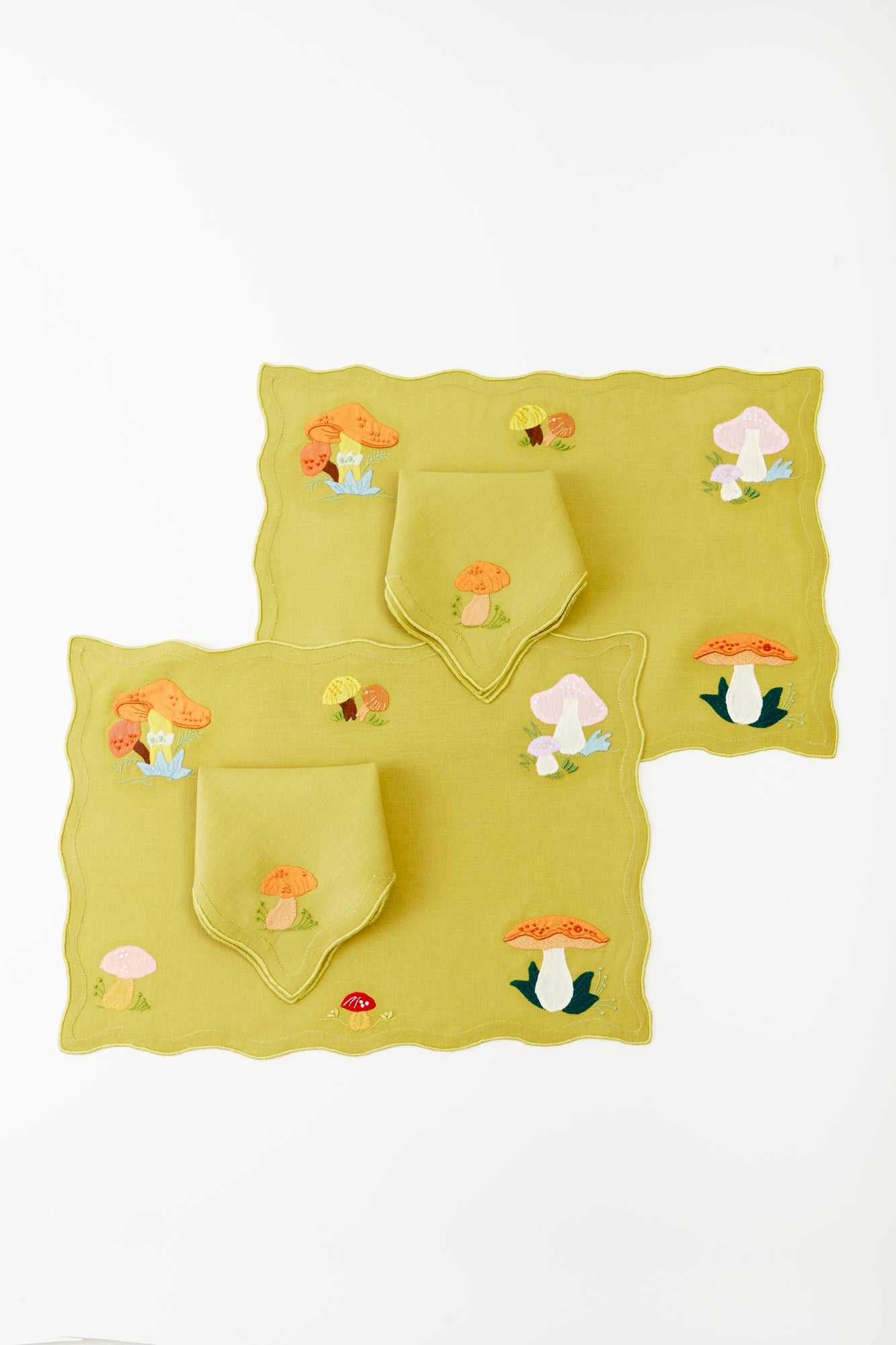 Set of two lime green linen placemats, each paired with a matching napkin, featuring embroidered mushrooms and botanical details in earthy tones. The scalloped edges and colorful mushroom motifs create a stylish, nature-inspired look, making this set a sophisticated yet playful addition to any tablescape.