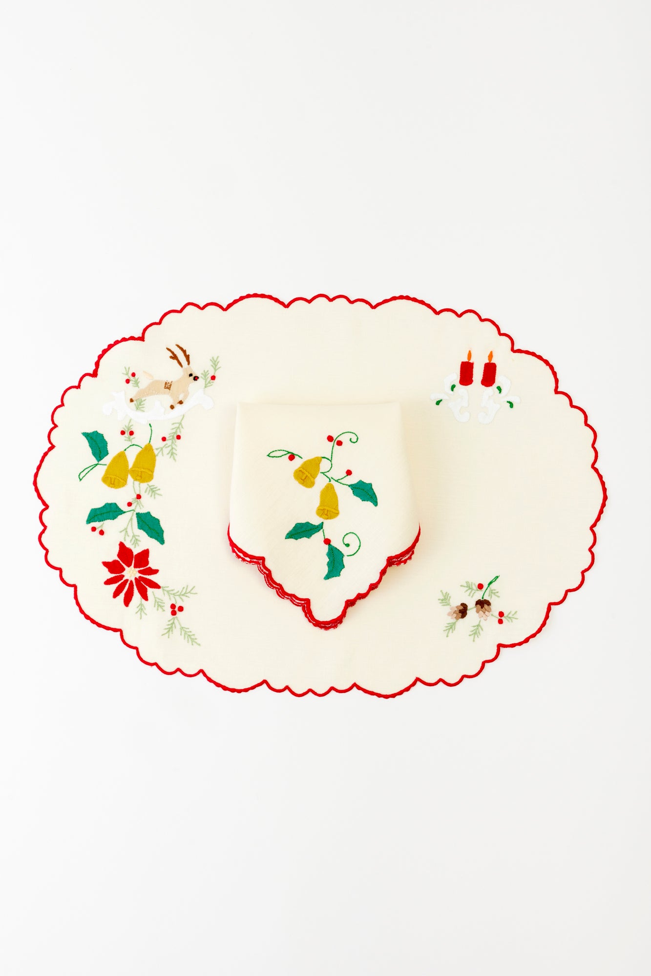 Ivory embroidered holiday tablecloth set with matching napkin, adorned with festive motifs including a reindeer, golden bells, red candles, and holly sprigs. The red scalloped edges enhance the traditional holiday aesthetic, creating a warm and inviting tablescape perfect for Christmas celebrations.