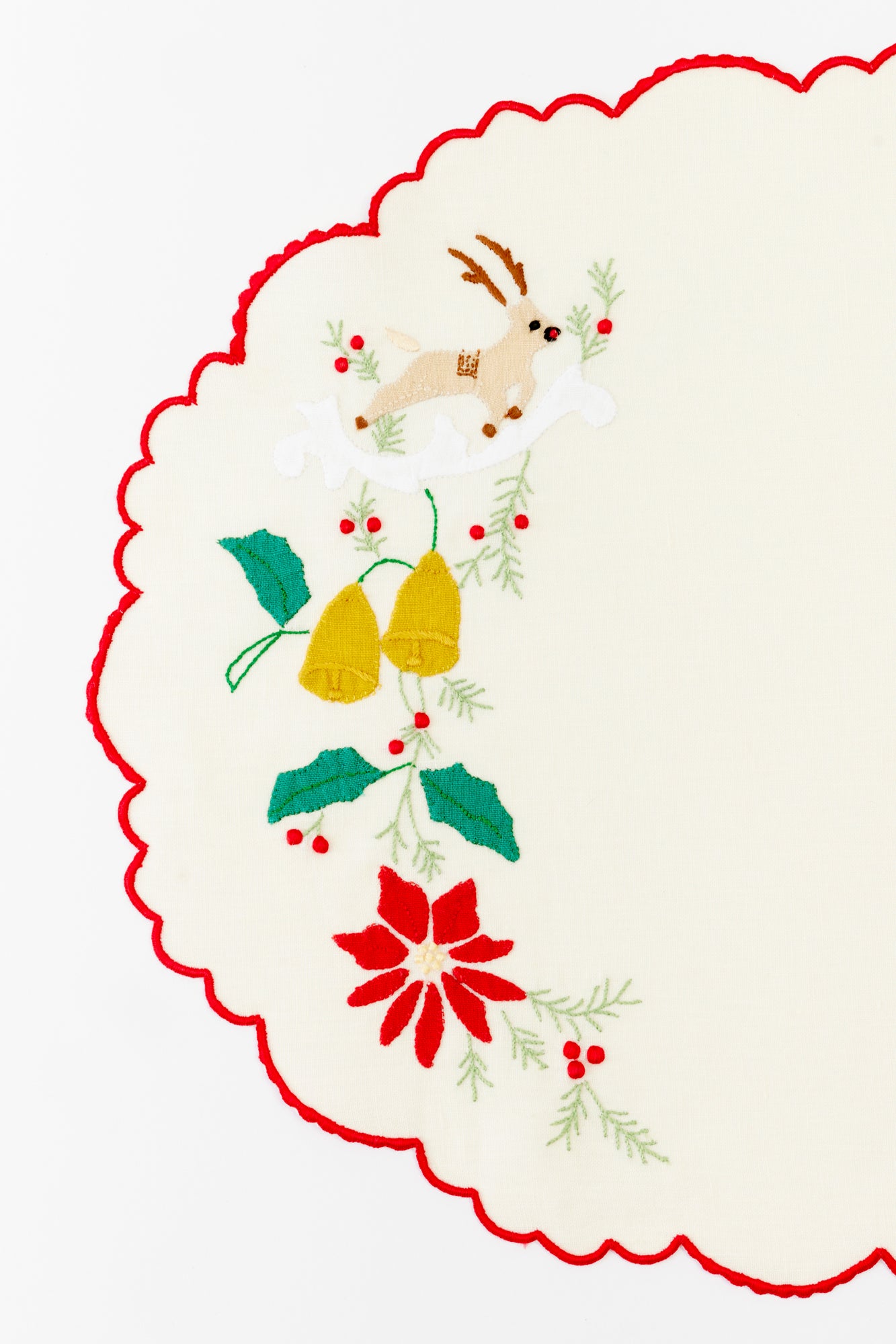 Close-up of an ivory holiday-themed tablecloth featuring intricate embroidery with a reindeer, golden bells, holly leaves, and a vibrant red poinsettia. The scalloped red border adds a festive and elegant touch, perfect for elevating a Christmas table setting with classic holiday charm.