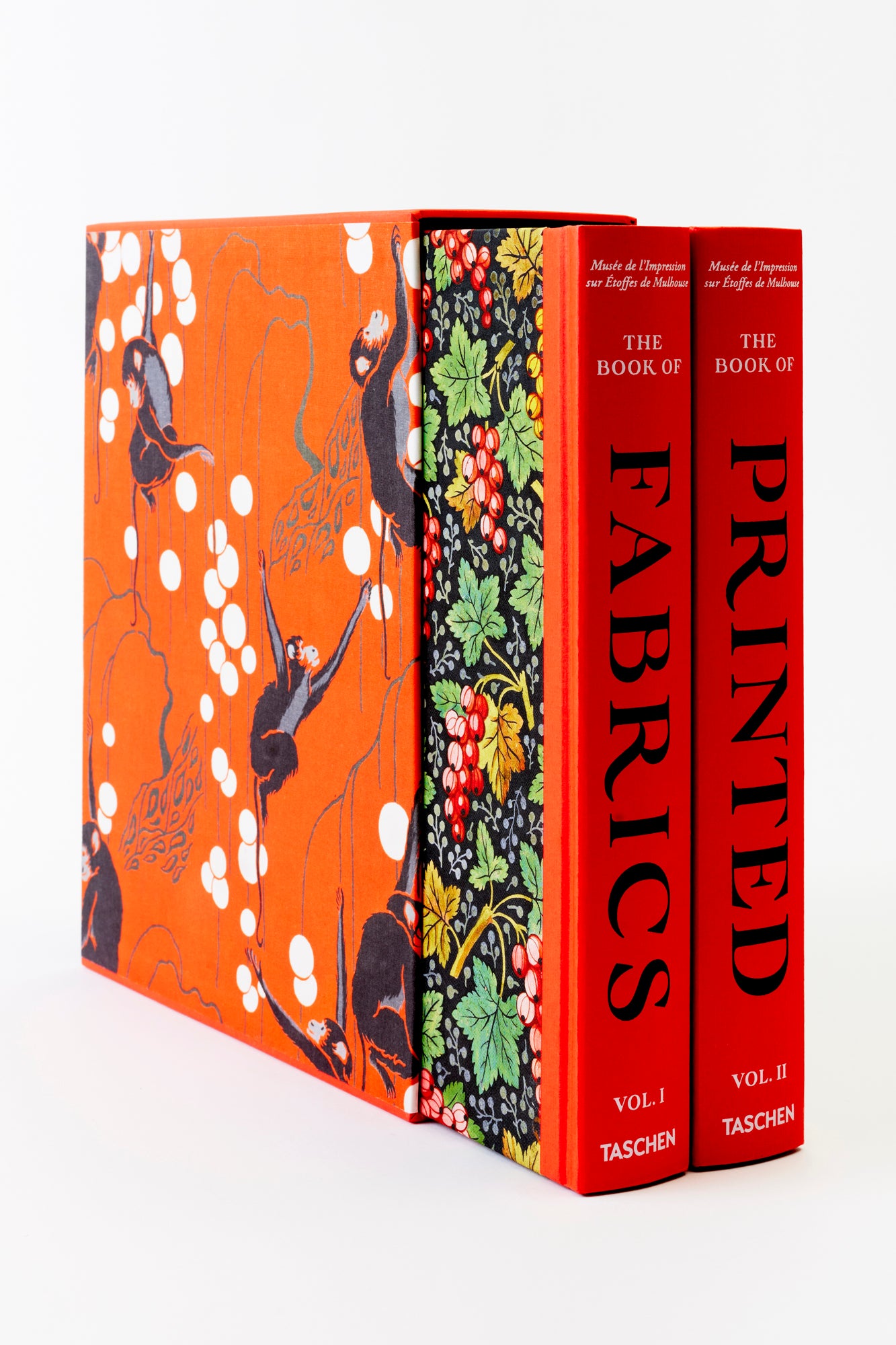 Close-up of two hardcover books, 'The Book of Fabrics' and 'The Book of Printed Fabrics,' enclosed in a vividly patterned slipcase. The books have bright red-orange spines, and the slipcase showcases an intricate design with floral motifs on a dark background.