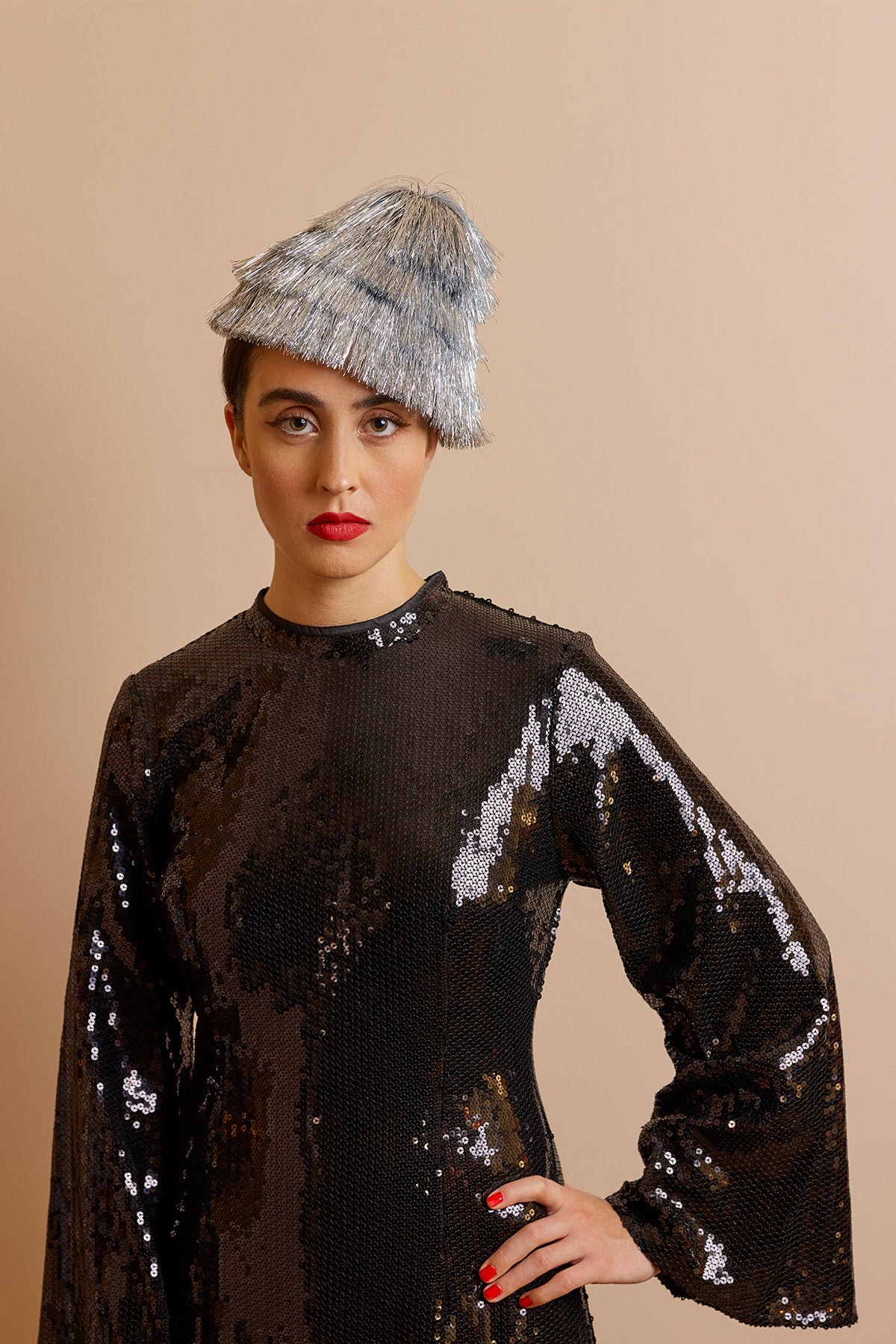 A person wearing a sequined black dress with bell sleeves and a green tinsel hat in the shape of a small Christmas tree. The background is a neutral tan, and the person has a bold red lip.