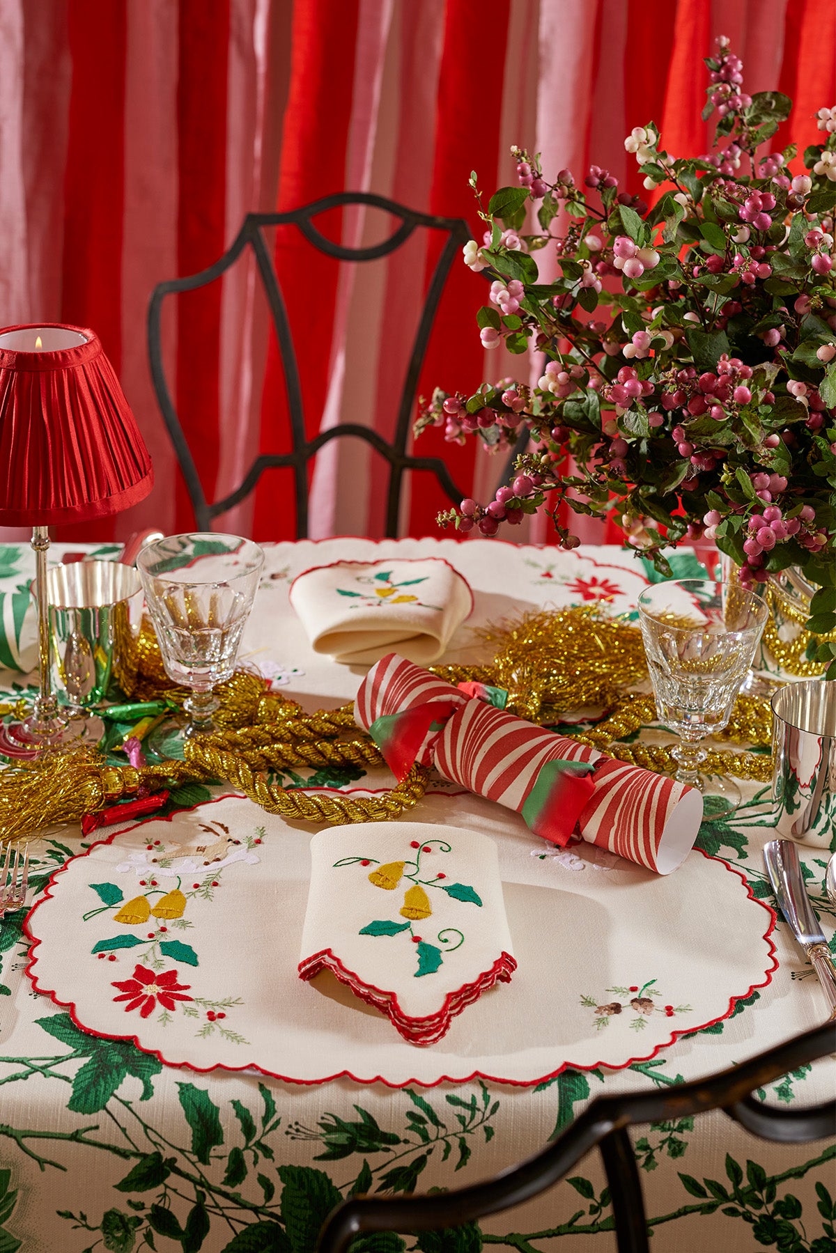 Elegant holiday tablescape with a classic red, green, and gold theme, featuring embroidered linens with festive bell and holly motifs. Crystal glassware, shiny gold tinsel, and a striped Christmas cracker bring a playful yet refined touch to this beautifully arranged table, set against a backdrop of pink and red curtains for a warm and inviting holiday feel.