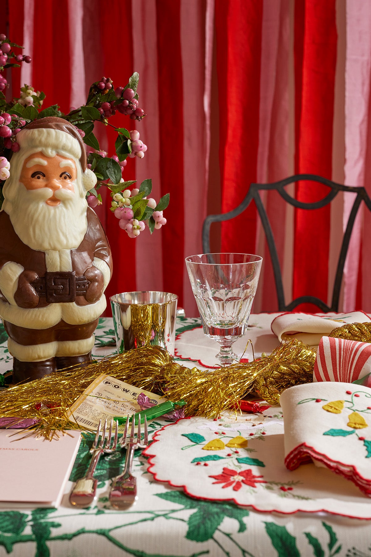 Festive Christmas table setting featuring a vintage-inspired chocolate Santa surrounded by sprigs of holiday berries. The table is adorned with crystal glassware, gold tinsel, and an embroidered holiday tablecloth with red and green details, creating a nostalgic yet chic holiday ambiance. A striped napkin and whimsical holiday décor complete the cozy and elegant scene.