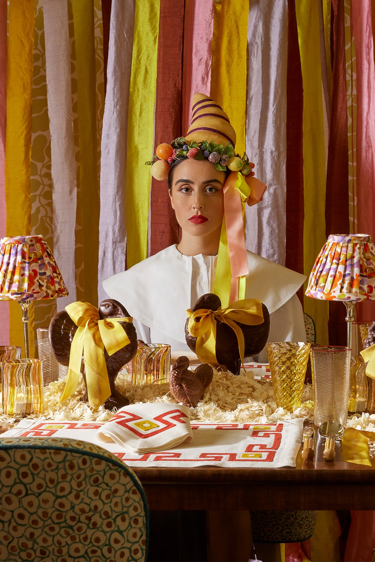 Striking holiday tablescape featuring a woman in a white, minimalist collared dress, adorned with a whimsical cornucopia hat topped with fruit-and-ribbon. She sits against a backdrop of colorful streamers, with chocolate turkeys tied in golden bows placed elegantly on the table. Amber-hued glassware and bold geometric napkins add a chic, playful contrast, completing the festive yet avant-garde aesthetic of this stylish Thanksgiving setting.