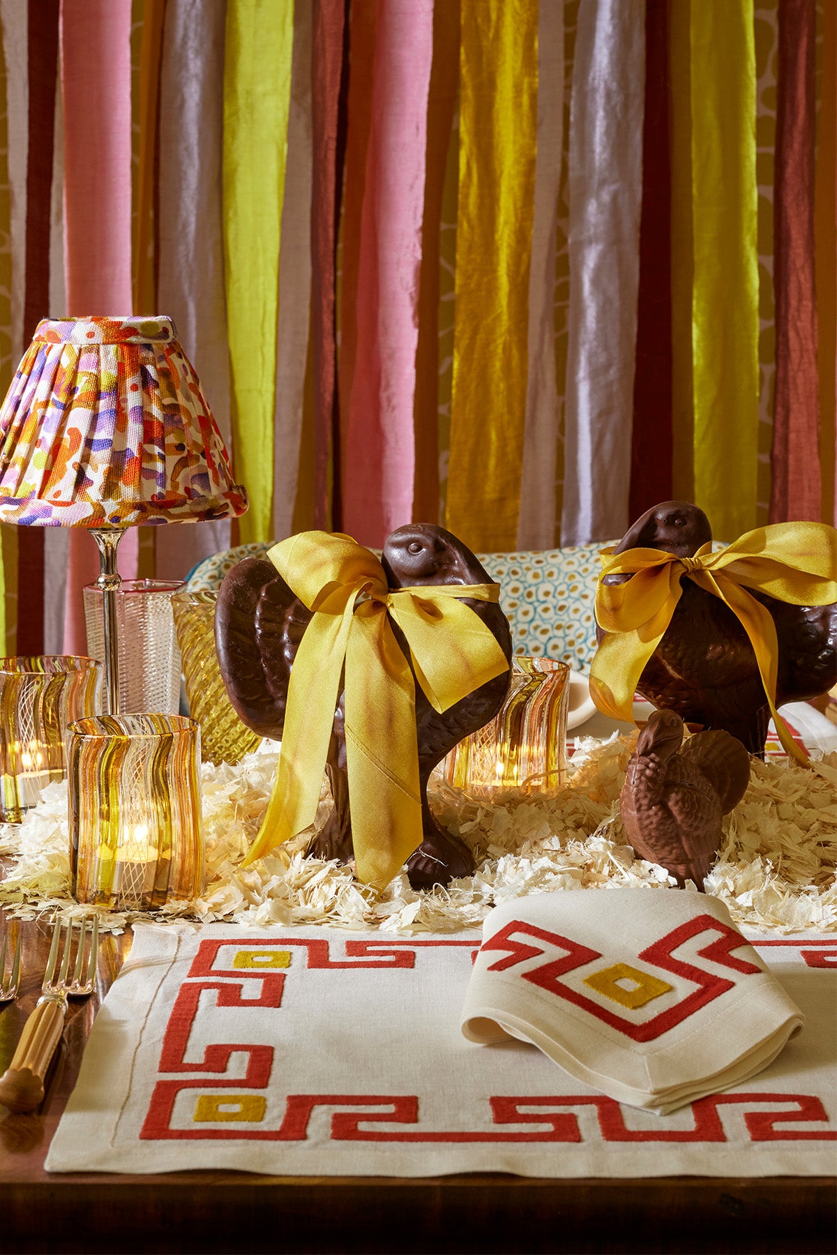 Elegant Thanksgiving table setting featuring chocolate turkeys adorned with golden satin bows, surrounded by amber glasses and colorful, textured napkins with bold geometric patterns. The scene is accentuated by playful, multicolored ribbon streamers in vibrant shades of yellow, pink, and red, creating a chic and festive ambiance perfect for a stylish holiday celebration.