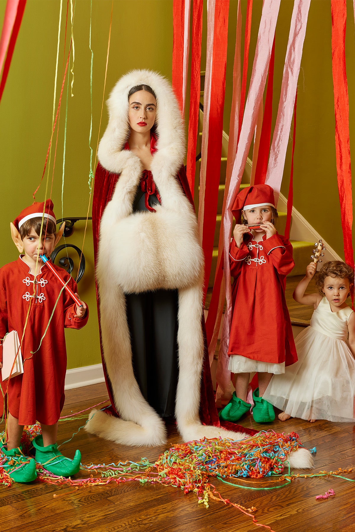 Elegant holiday cheer as a mother in a stunning fur-lined cape stands with her children dressed as Christmas elves, framed by cascading pink and red ribbons. A chic, festive moment full of whimsy and style.