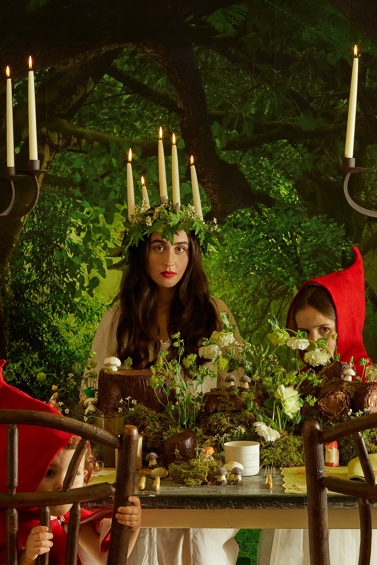 A captivating, ethereal tablescape set in a lush, forest-like atmosphere, where a woman in a white cottagecore inspired dress wears a brass crown adorned with flowers, ivy and taper candles. Surrounded by moss, chocolate mushrooms, and tiny woodland figurines, the scene evokes a mystical forest feast, with a lime green placemat adding a playful touch to the rustic charm.