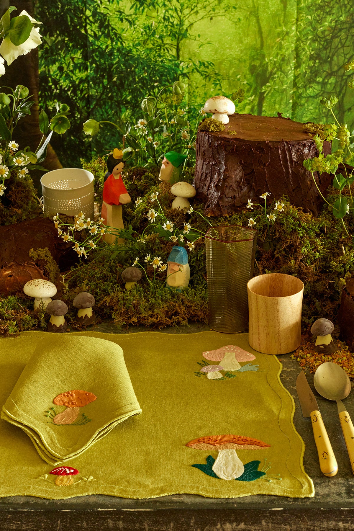 A whimsical woodland-themed table setting featuring a moss-covered landscape with small wooden figurines of Snow White and the Seven Dwarfs, delicate mushrooms, and fresh white wildflowers. A lime green embroidered placemat with mushroom designs adds a touch of nature-inspired charm, while earthy wooden cups and glassware complete this enchanting forest scene.