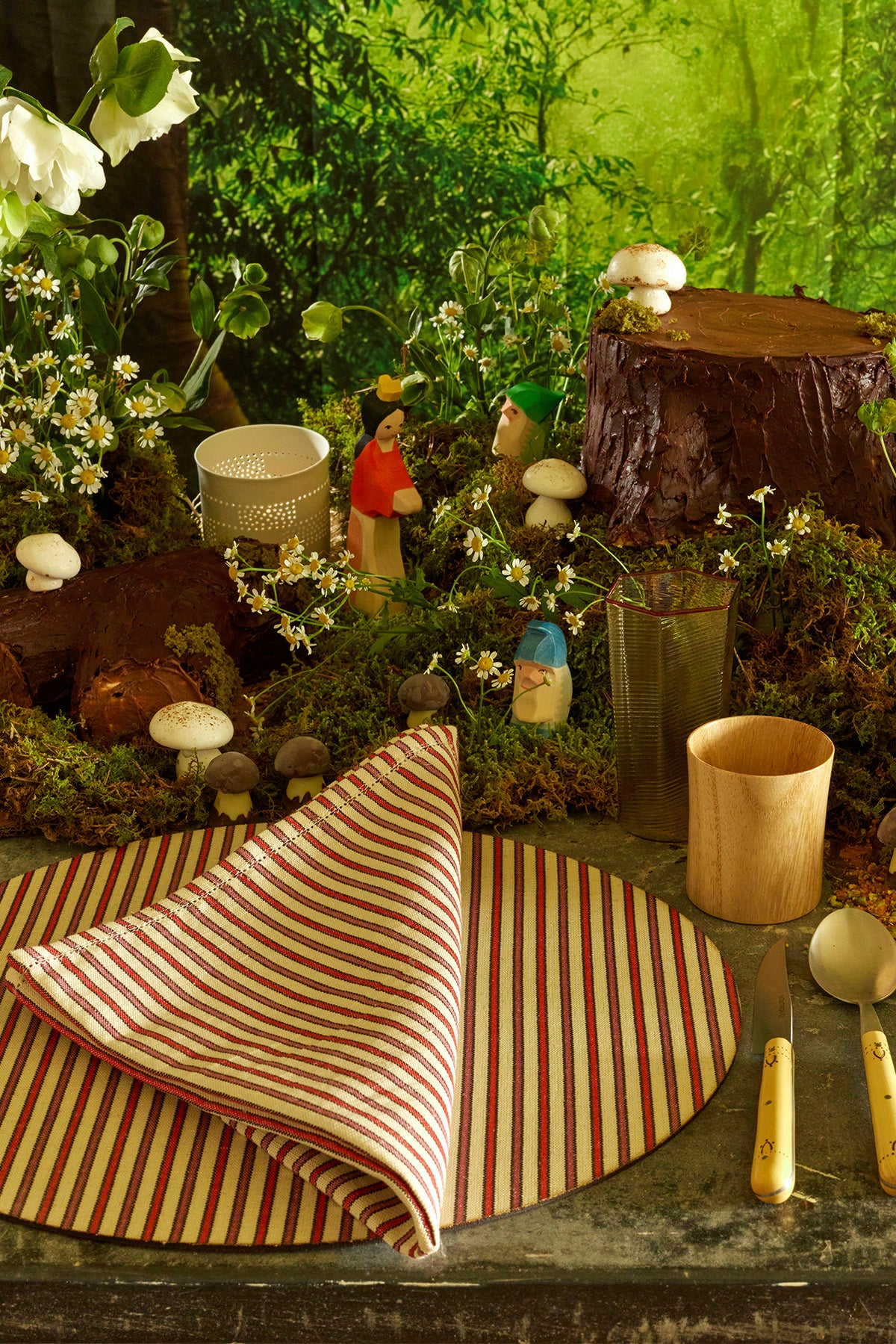 The image shows a whimsical, woodland-themed table setting. At the center, there is a round striped placemat with a folded napkin in red and white stripes, alongside vintage-style cream-colored cutlery and a wooden cup. The background features moss, small mushrooms, and figurines of a fairy-tale character and a gnome, all arranged around faux tree stumps and white flowers. The lush, green forest backdrop completes the magical, nature-inspired scene.