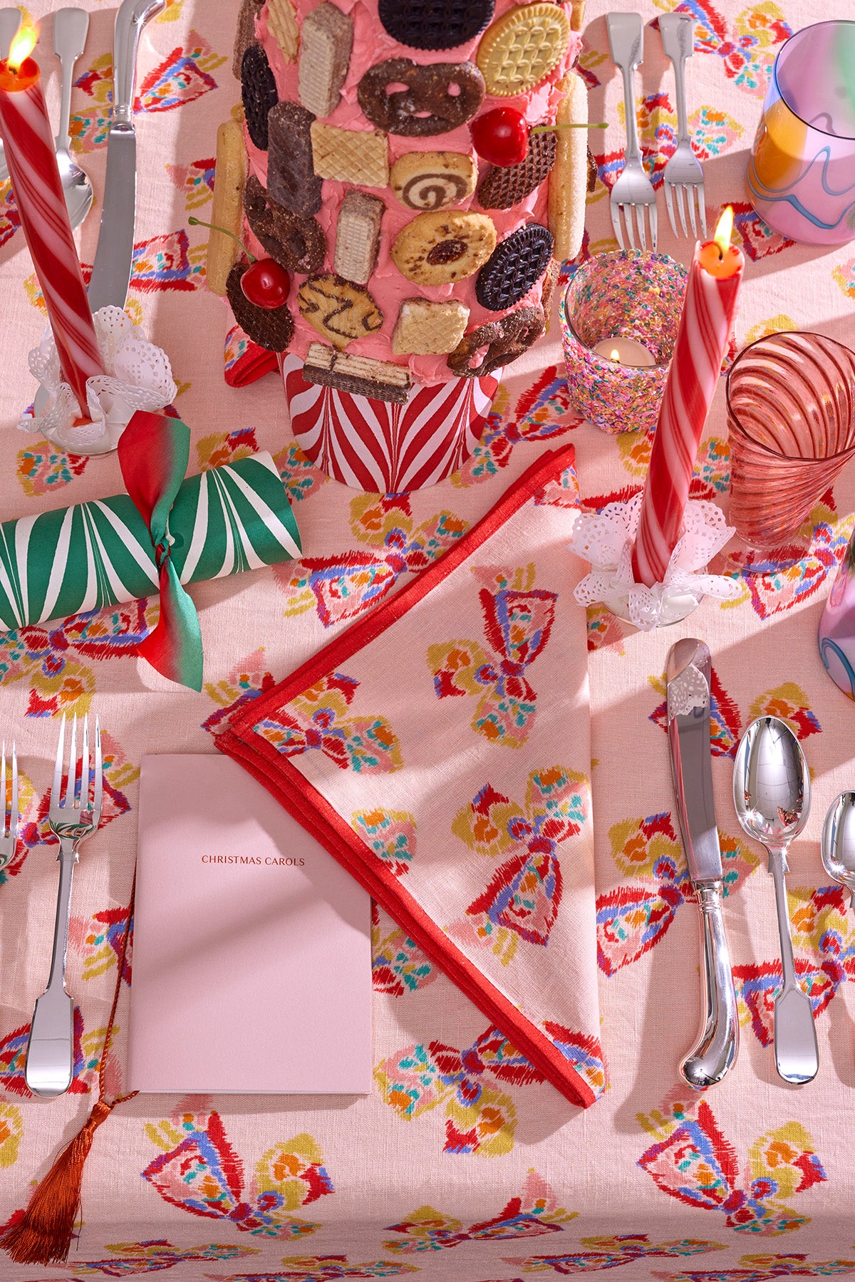 Festive and whimsical holiday table setting featuring a towering cookie display with colorful pink frosting and decorated with an array of wafers and biscuits. The table is adorned with vibrant patterned linens, pepperming candy-striped candles, and a green and red Christmas crackers. A pastel pink booklet labeled 'Christmas Carols' adds a charming touch, while a multicolored tumbler and sequin-adorned votive holder complete the playful, candy-inspired aesthetic.