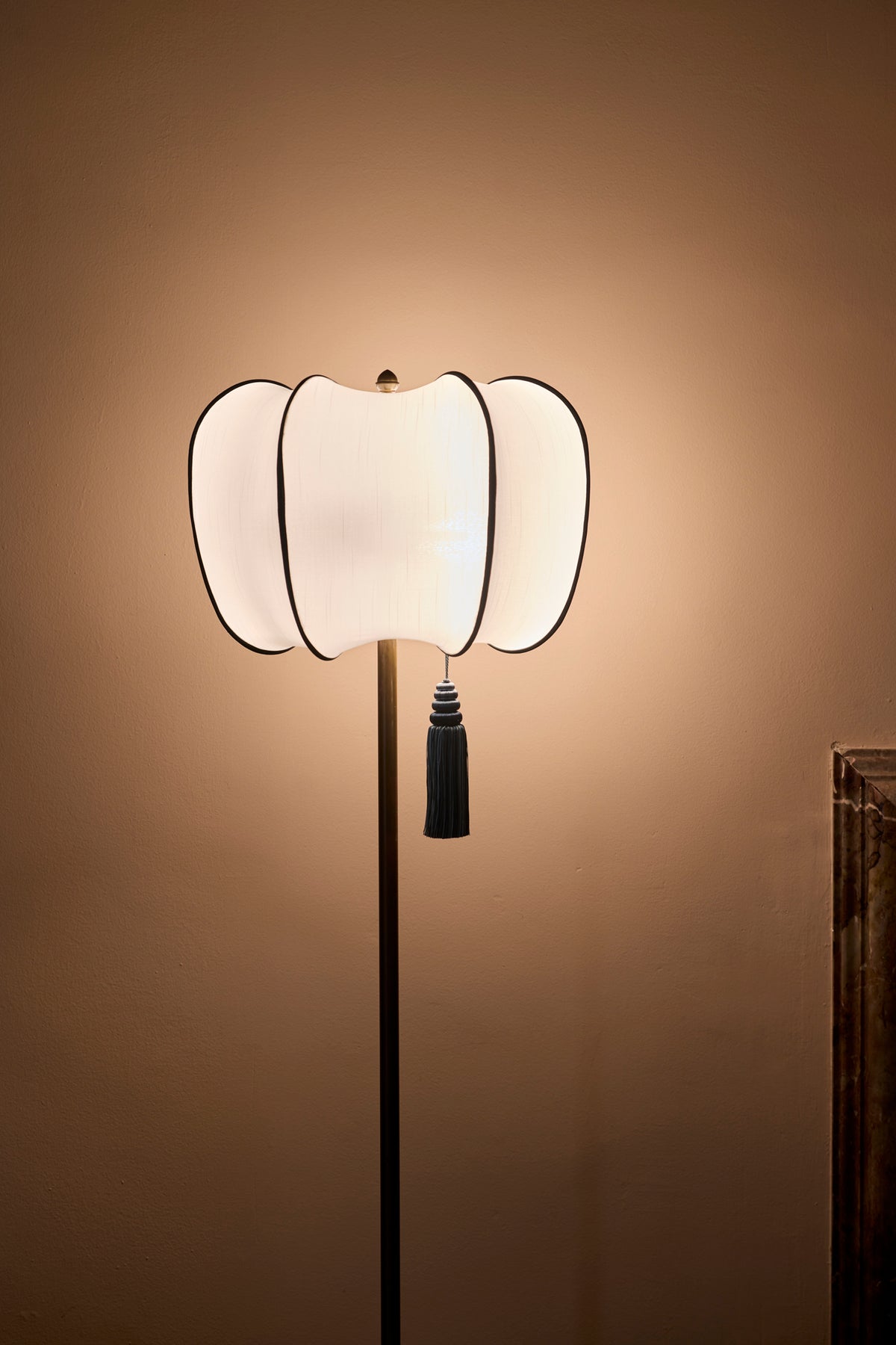 A modern floor lamp with a sculptural, ribbed white shade outlined in green, featuring a black tassel and a slim black stand, set against a warm brown wall.