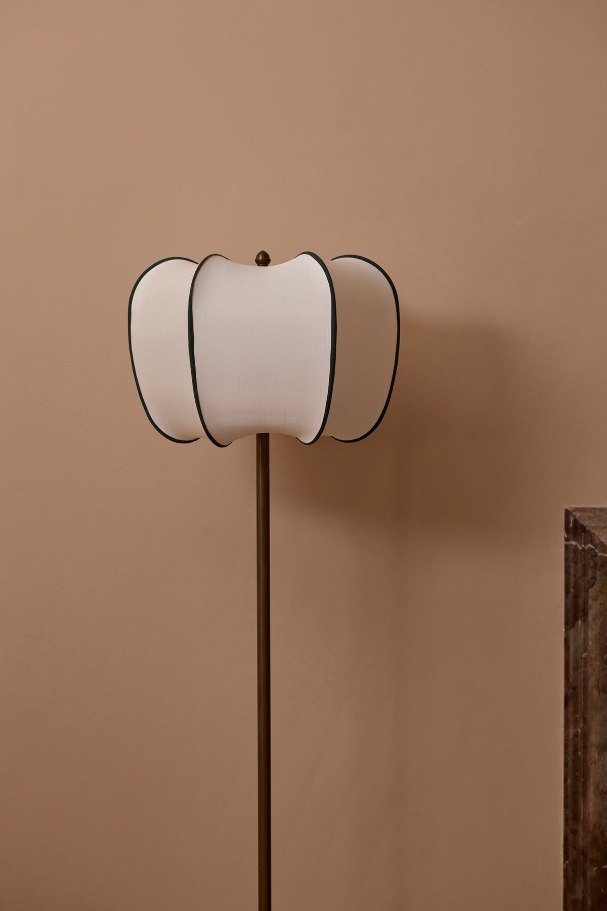 A modern floor lamp with a fluted, wavy white shade outlined in green, mounted on a slim dark metal pole, set against a solid warm beige wall.
