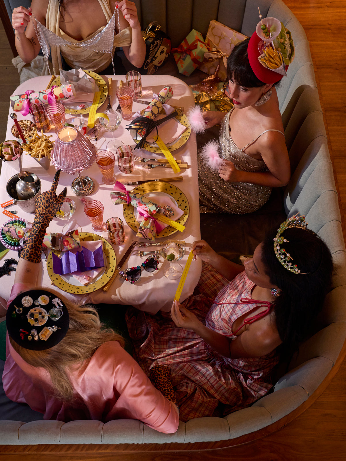 Four women seated around a decorated dining table in a curved upholstered booth, wearing colorful party hats and playful headbands, with drinks, plates, and wrapped gifts on the table.
