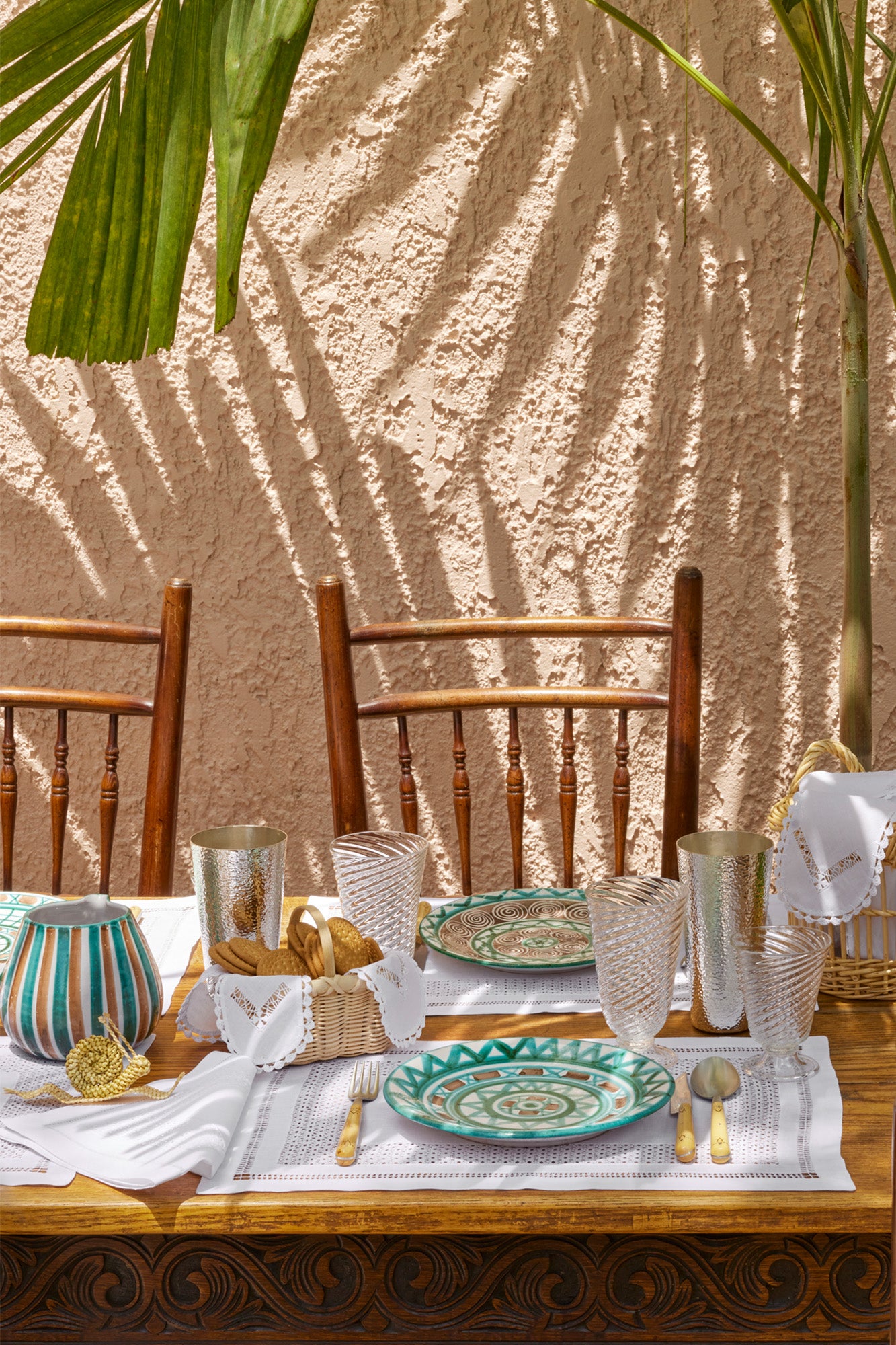 Table setting under a palm frond. Wooden table with carved details, white linen placemats, blue patterned plates, wicker breadbaskets with openwork embroidered linen fabrics, hand blown glass goblets, and a corn husk snail. 