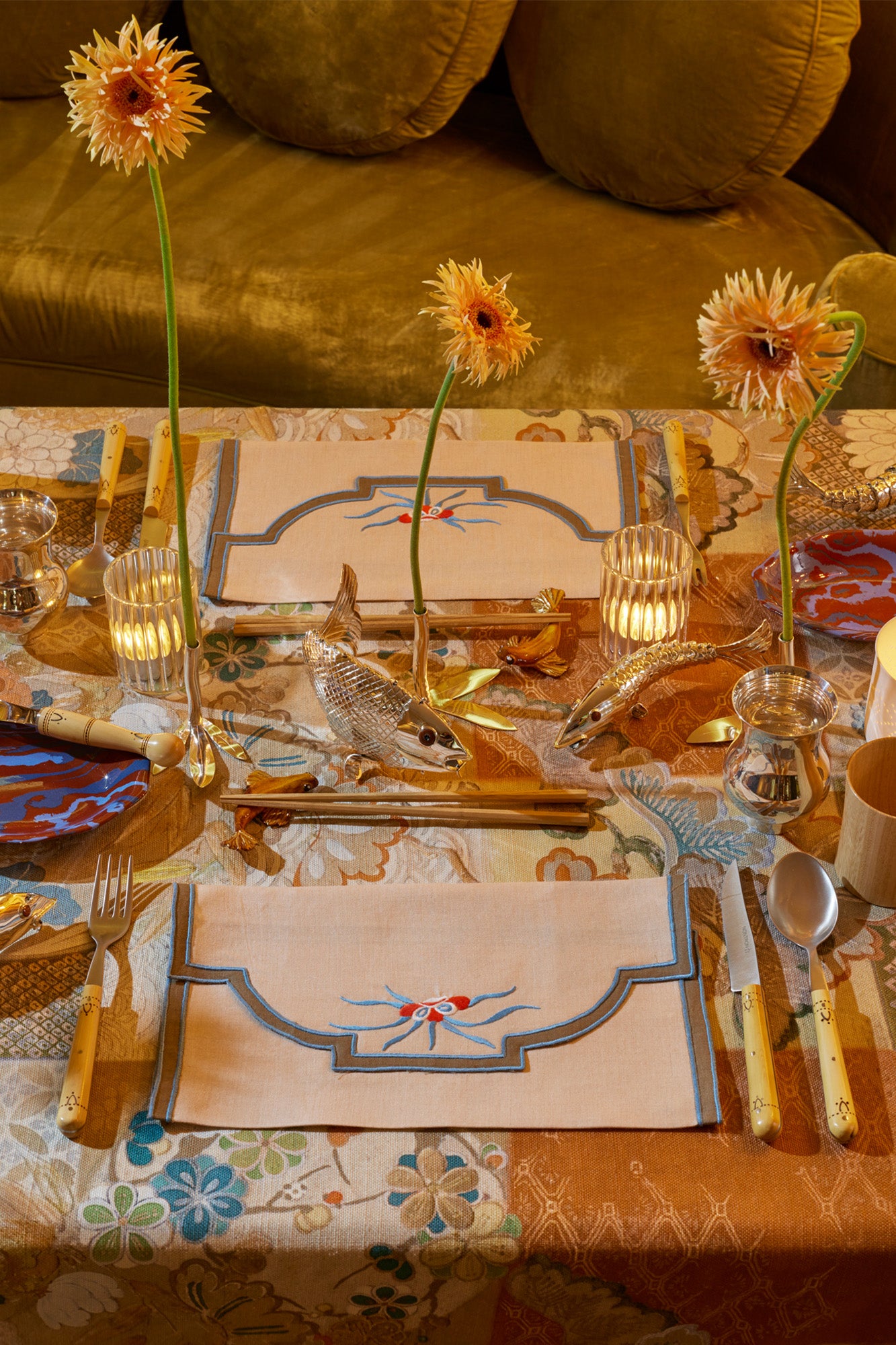 Table setting for two people. Tan placemats set on top multicolor tablecloth. Boxwood silverware and wooden chopsticks. Chopsticks sit on fish shaped chopstick rests. Silver goblets and marbled ceramic dishes. Silver articulated fish and fresh yellow flowers with long stems. Glass votives with tea lights inside. 