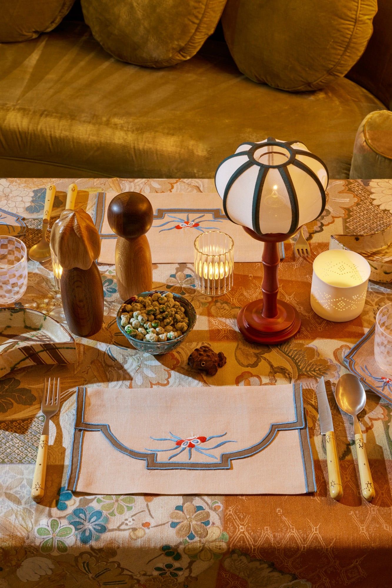 Close up photograph of table setting featuring embroidered linen placemats, boxwood silverware, a table lamp with a white shade, green trim, and red base, wooden salt and pepper grinders, fluted glass votive holders, all atop a multicolor tablecloth. 