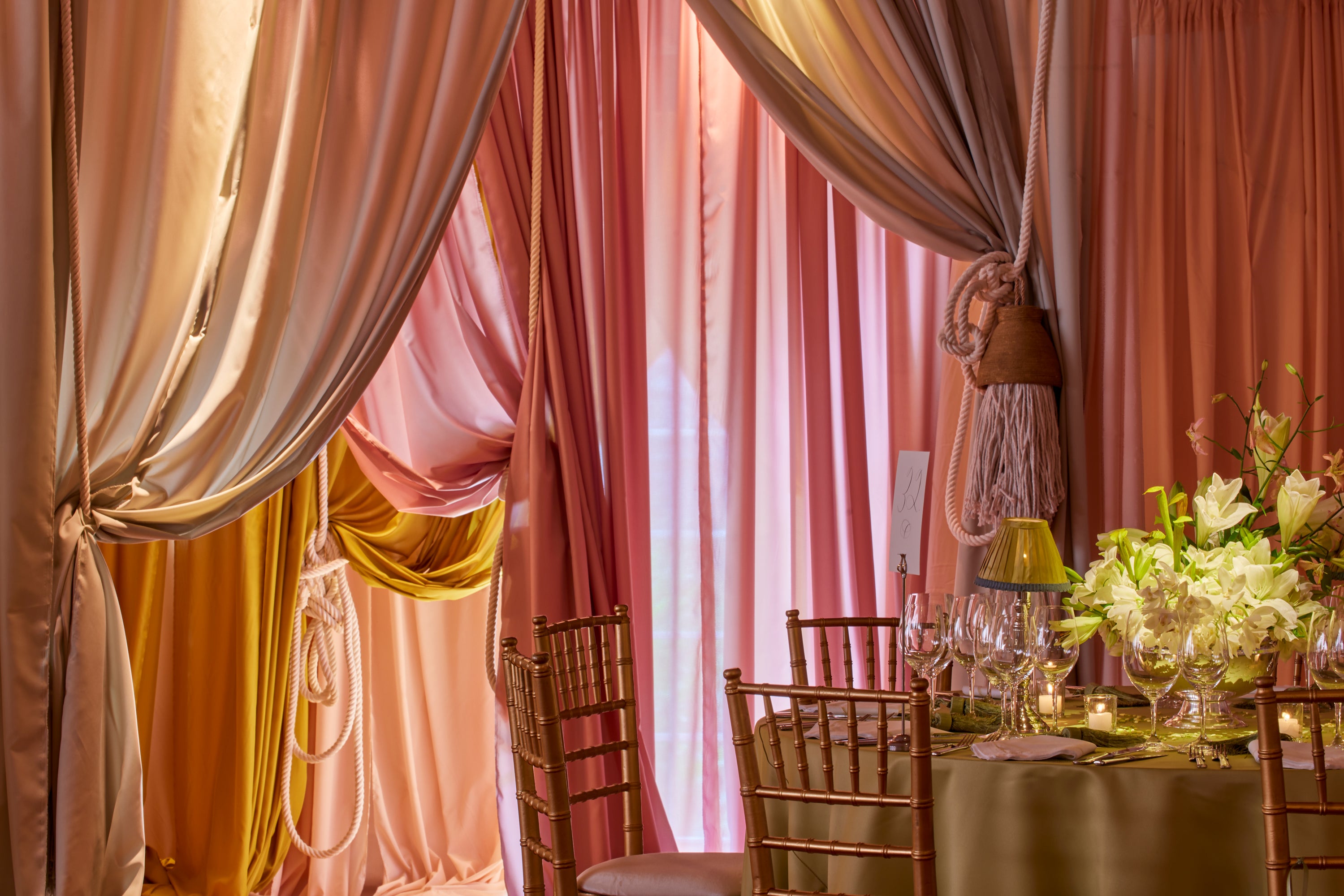 Pink and orange drapery behind a table set with elegant white lilies, green table lamps, and wine glasses on a green tablecloth