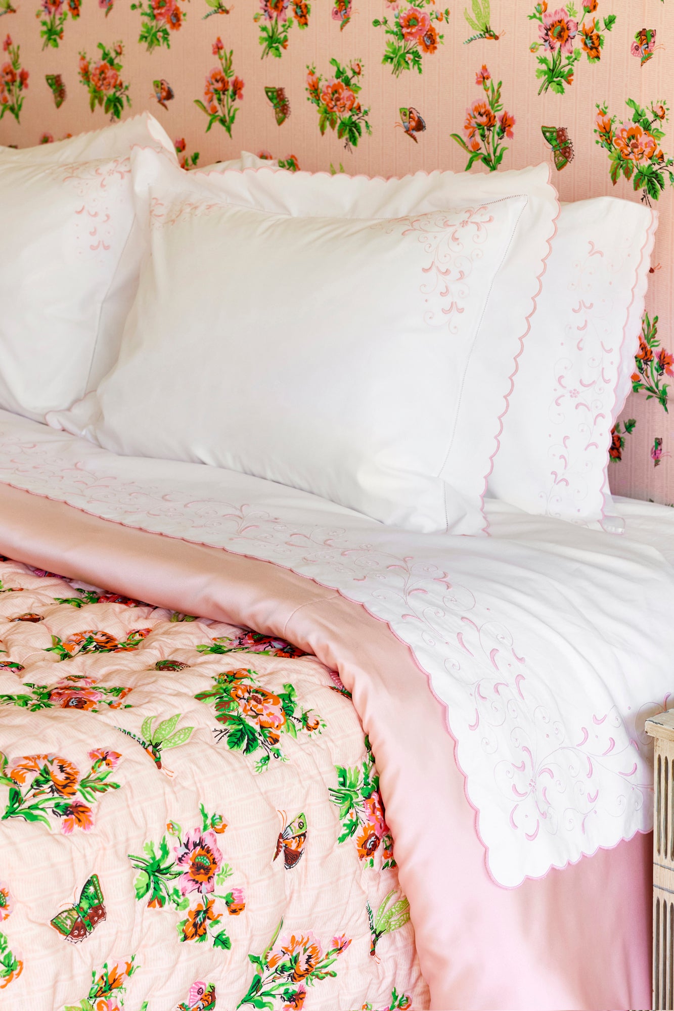 Bed featuring white percale sheets with pink embroidery, quilted floral coverlet, and headboard against a floral wallpapered wall.