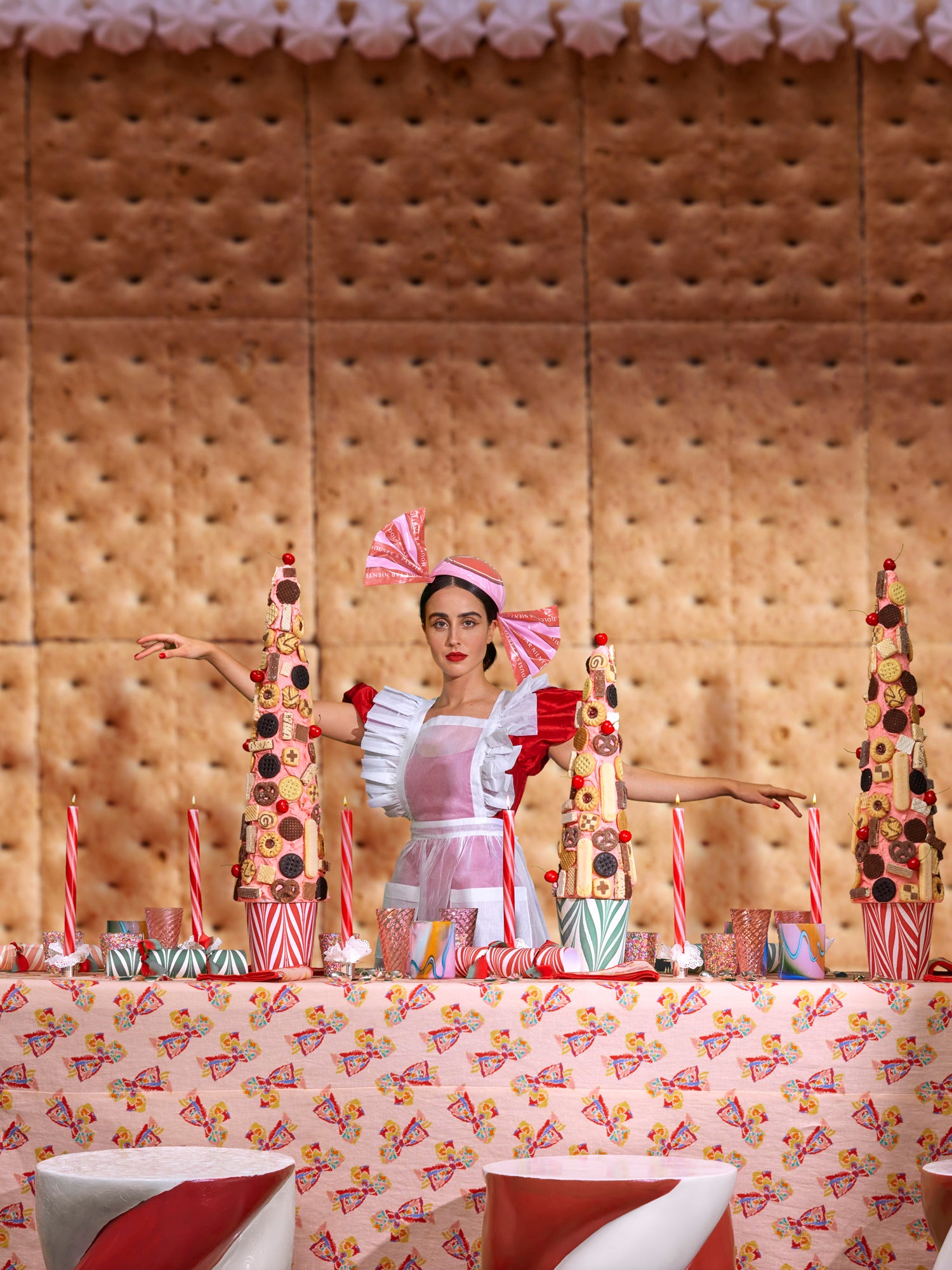 Bold, theatrical dessert table scene with a woman dressed in a playful, candy-striped bow headpiece and a red and white apron, standing between two towering cookie displays. The festive table is decorated with vibrant, bow-patterned linens, peppermint striped candles, and pastel-colored glassware, creating a whimsical, candyland-inspired aesthetic.