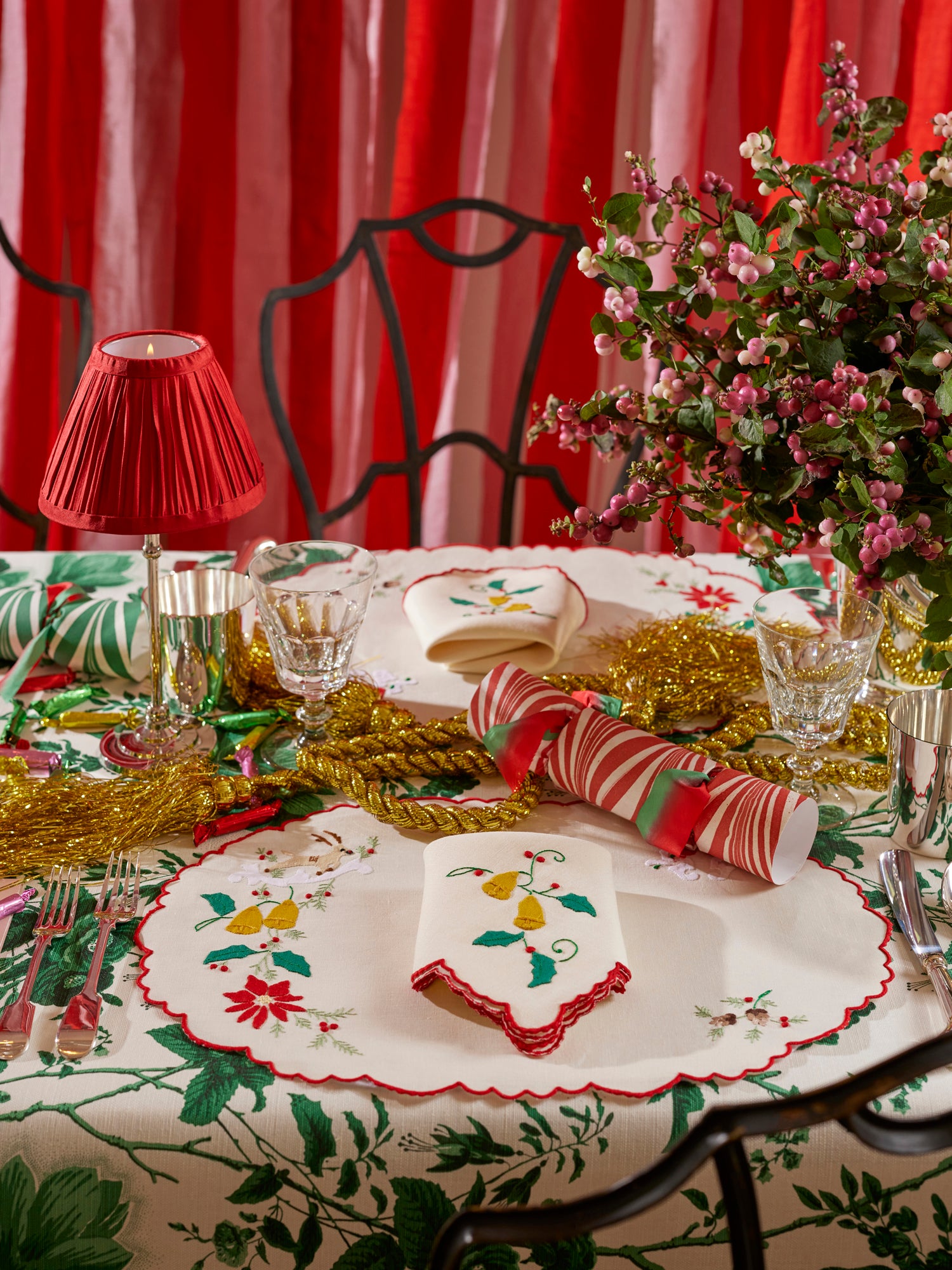 Christmas themed table setting featuring silver tumbler, crystal goblet, silver table lamp with red shade, linen placemat and napkin with French embroidered appliqué, fresh florals, and marbled paper party crackers. 