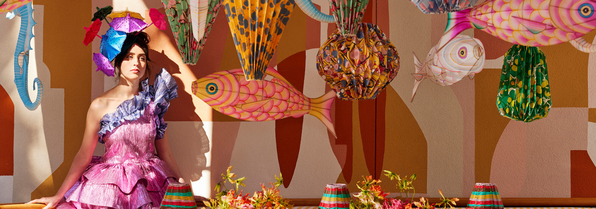 A person in a pink ruffled dress against a wall adorned with colorful paper fish lanterns and marbled paper lanterns. The vibrant scene includes striped lampshades and glass vases on a table, creating a whimsical, artistic atmosphere.