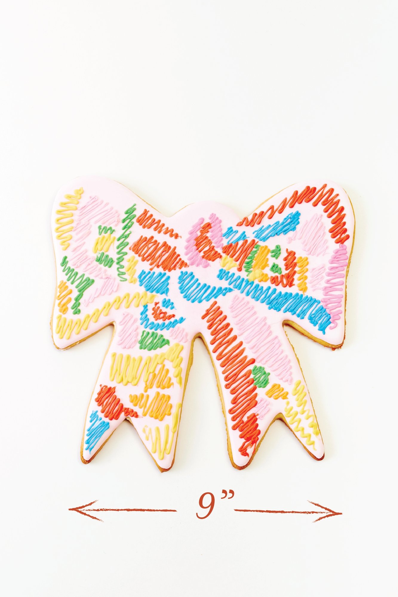 A colorful bow-shaped cookie with a vibrant, hand-drawn look. The cookie is covered in pink icing with an abstract pattern of multicolored squiggly lines, creating a bold and unique design.