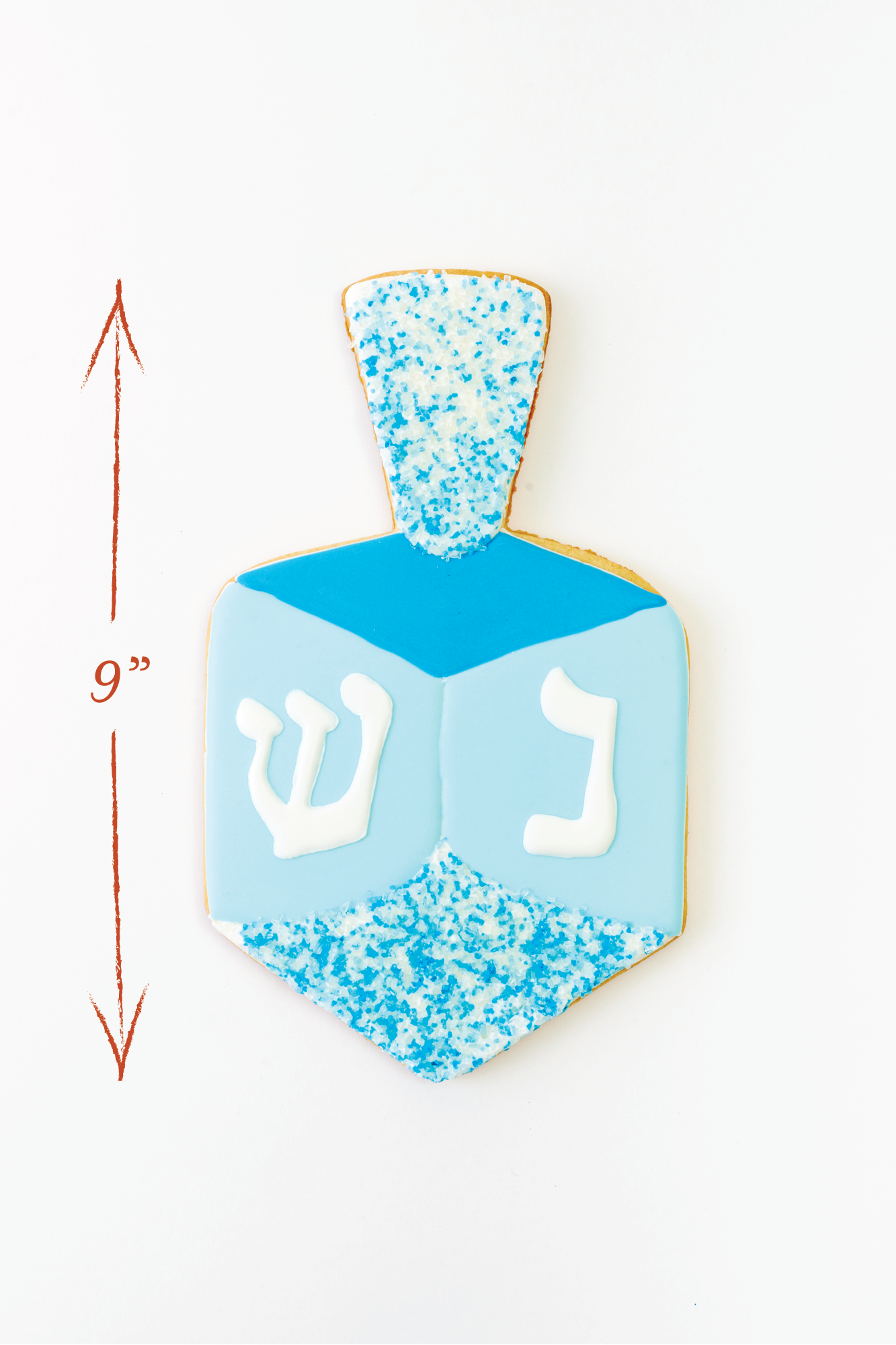 A dreidel-shaped cookie decorated in blue and white icing with sugar crystals at the top. The sides feature Hebrew letters in white, bringing a festive touch to this Hanukkah-inspired treat.