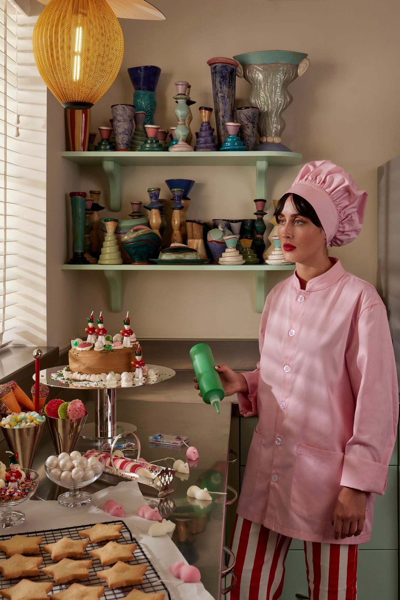 Person in a pink chef's outfit holding a green bottle in a kitchen setting with shelves of decorative items.
