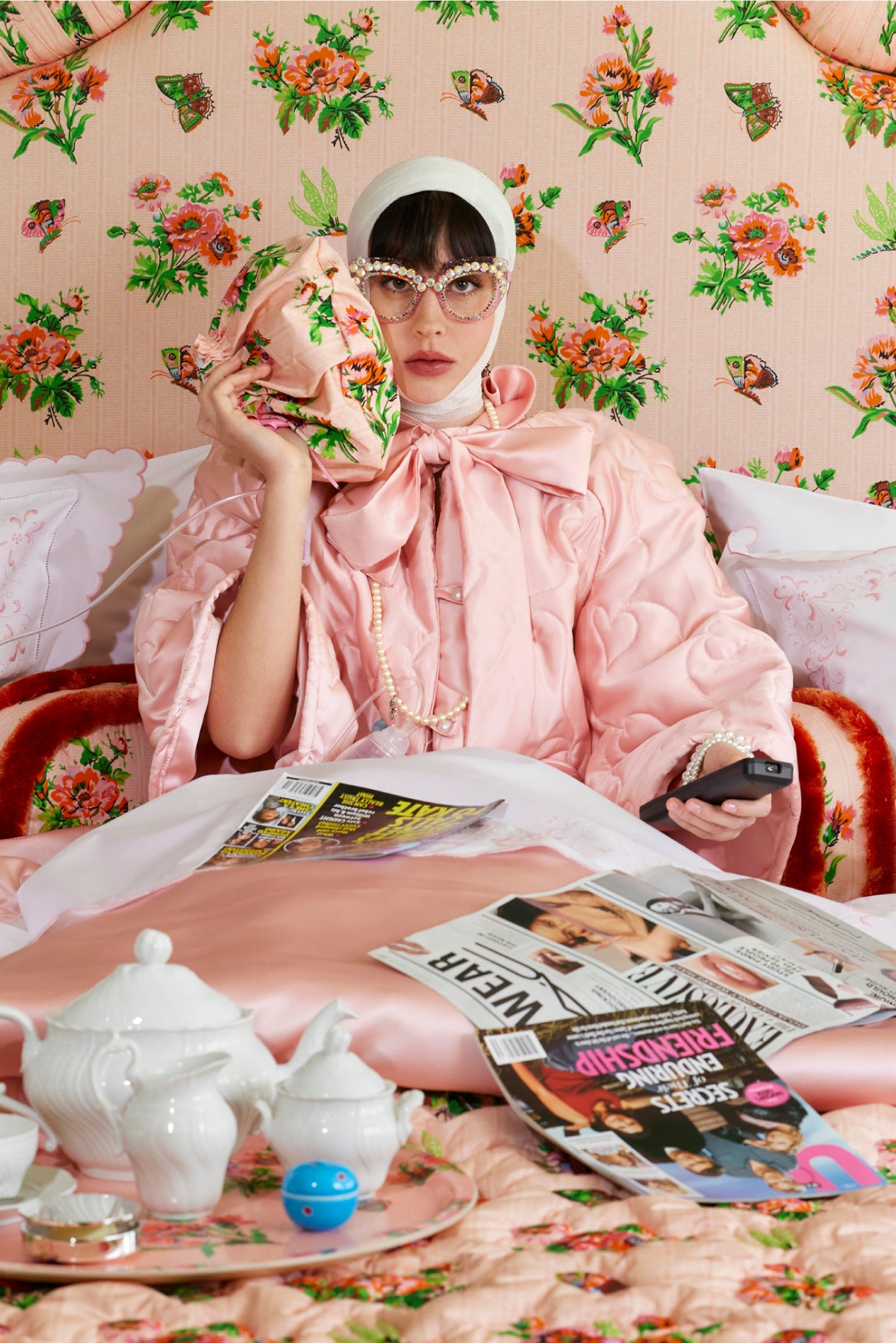 Woman in a pink quilted bed coat against a floral chintz headboard, sitting on a bed with a pink floral-patterned ice pack cover.