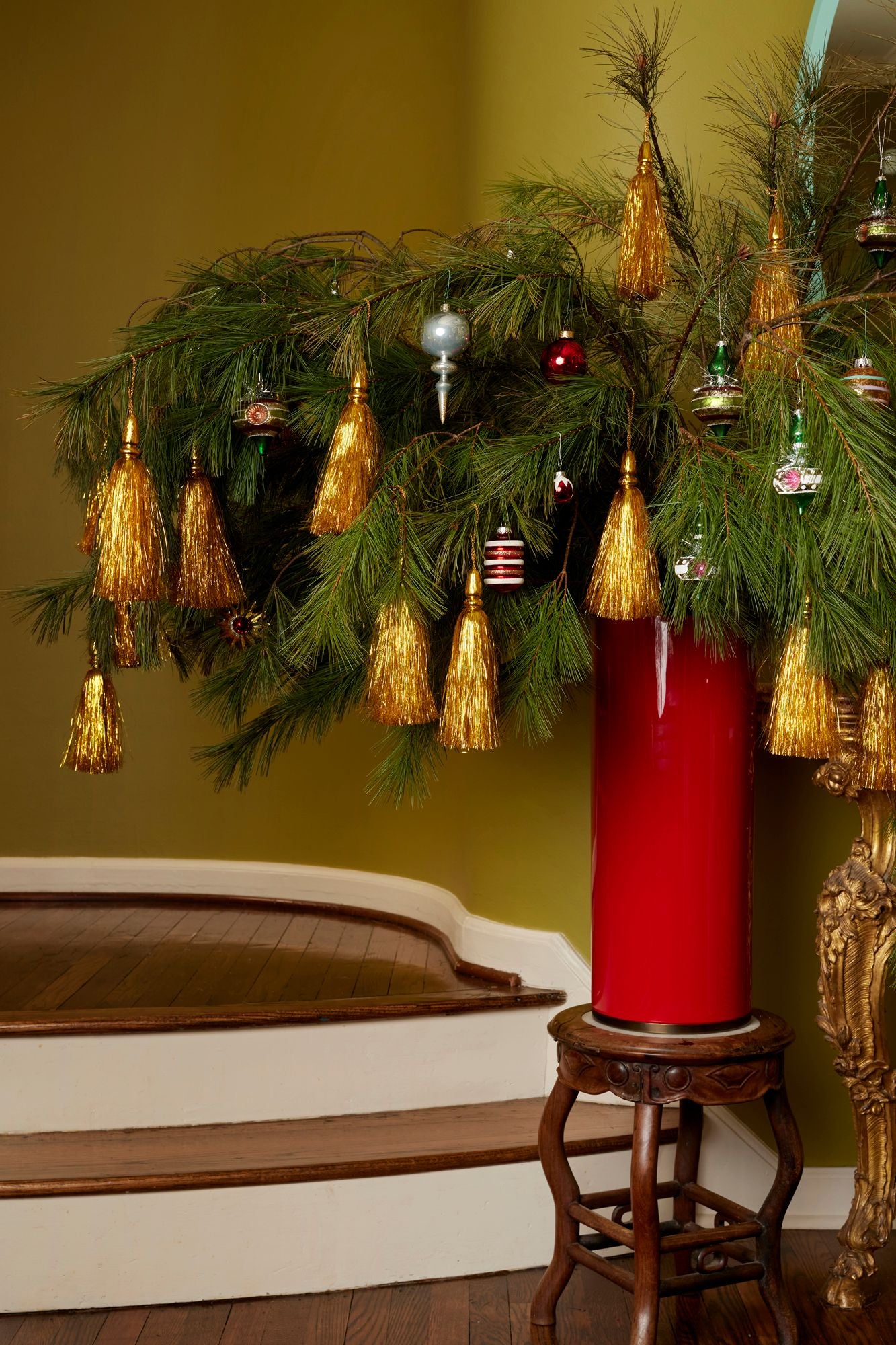 Decorative arrangement with greenery, ornaments, and a red umbrella vase on a wooden stool against a green wall.
