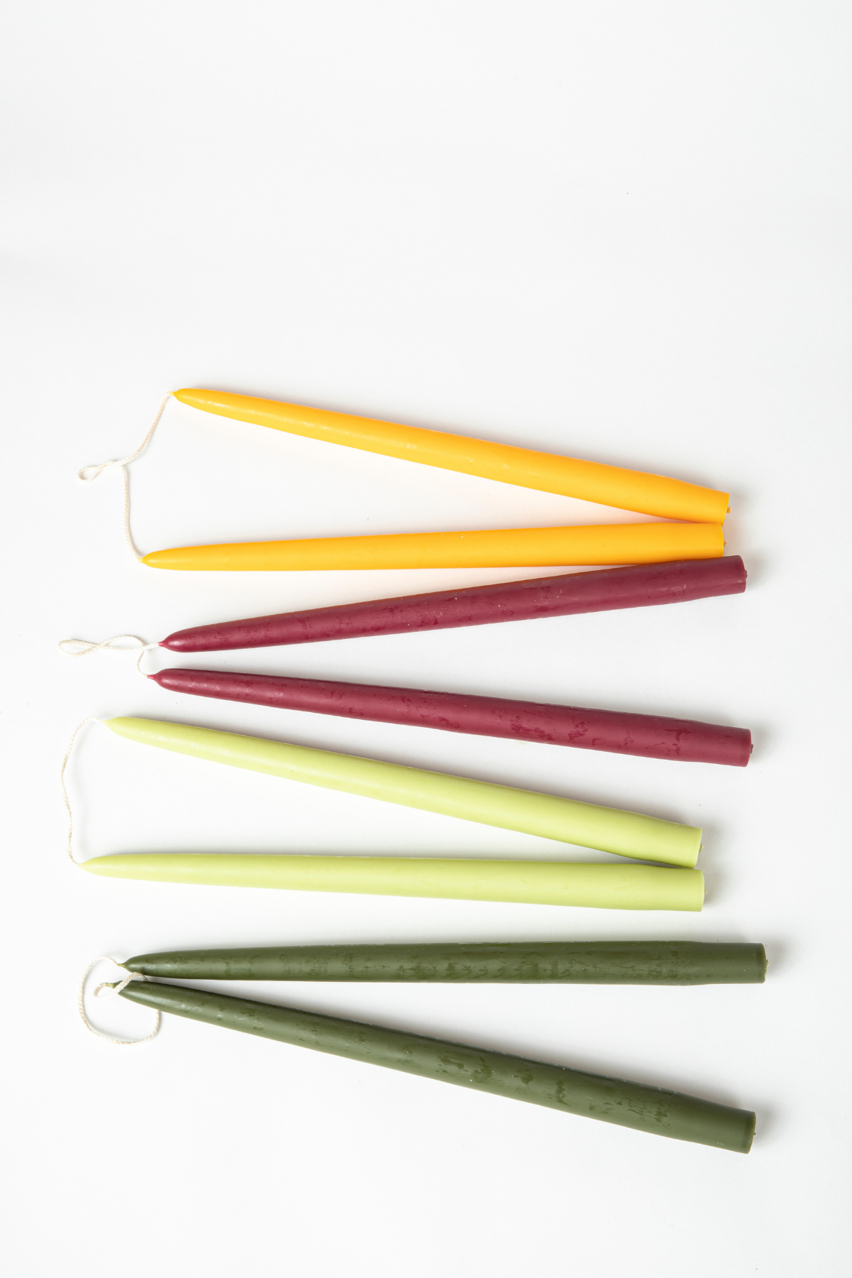 4 pairs of taper candles resting on a white background. Candle sets are yellow, maroon, light green, and dark green.