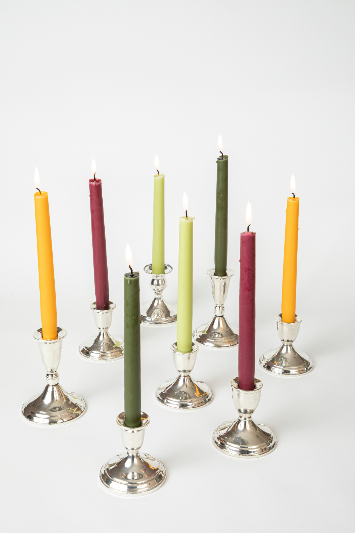 8 lit taper candles standing in silver holders against white background. Candles are yellow, maroon, light green, and dark green. 