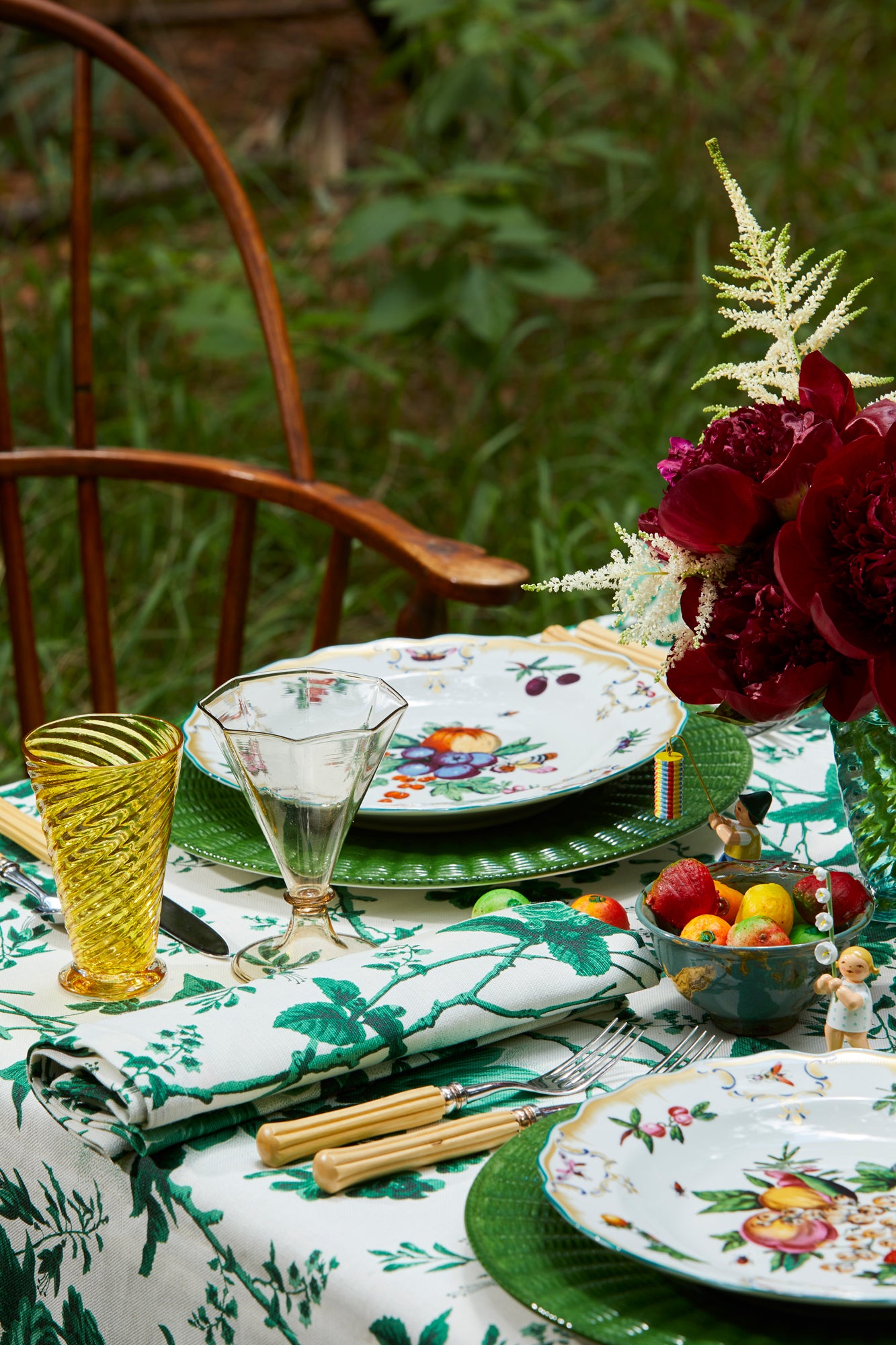 Photograph of a table setting featuring hand blown glass goblets, "De La Tour" green and white floral printed tablecloth and napkins, fluted boxwood silverware, porcelain plates and chargers, and fresh flowers. 