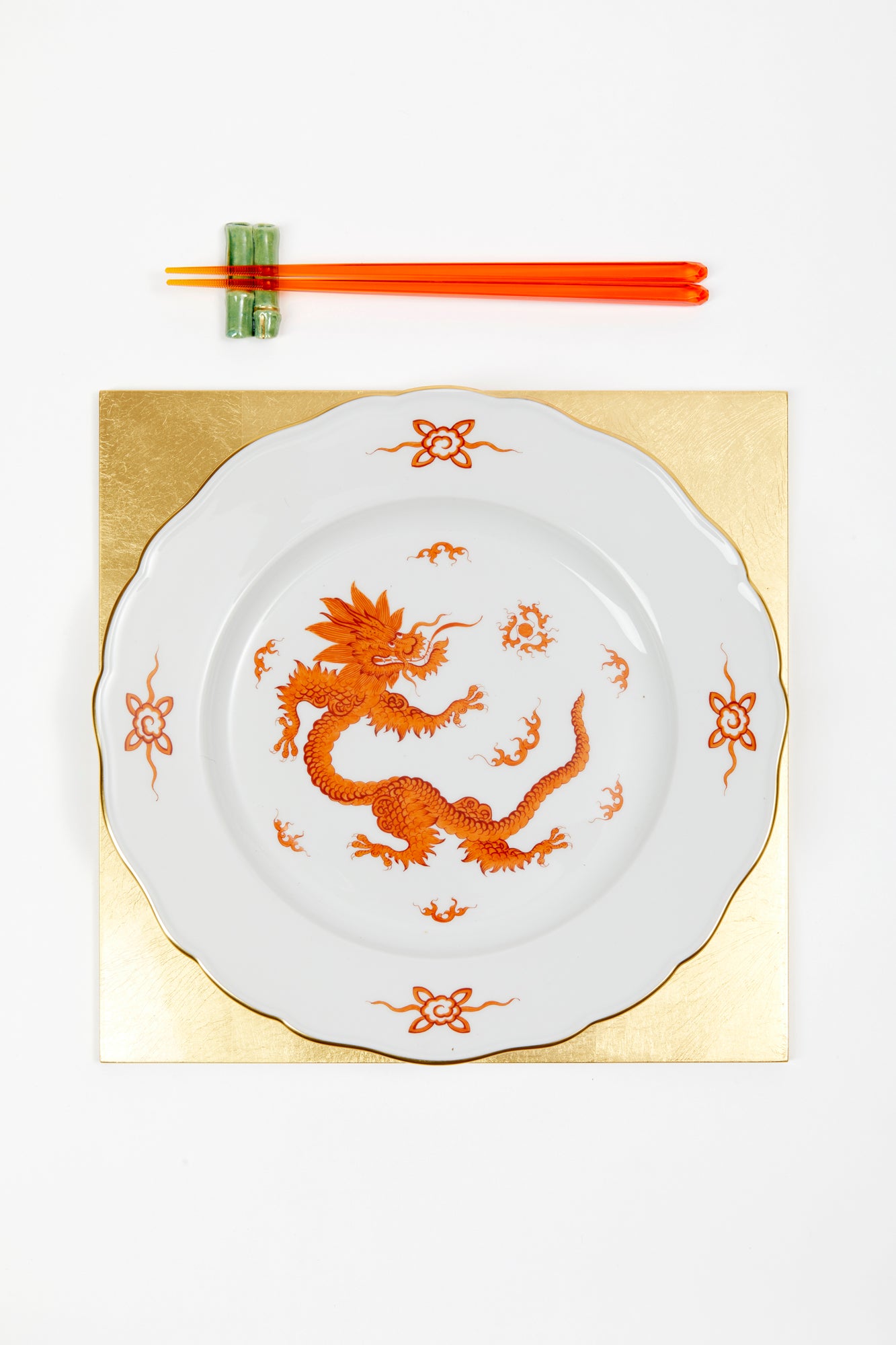 Photograph of table setting featuring square gold placemat, porcelain dragon dinner plate, and red chopsticks on porcelain bamboo rests.