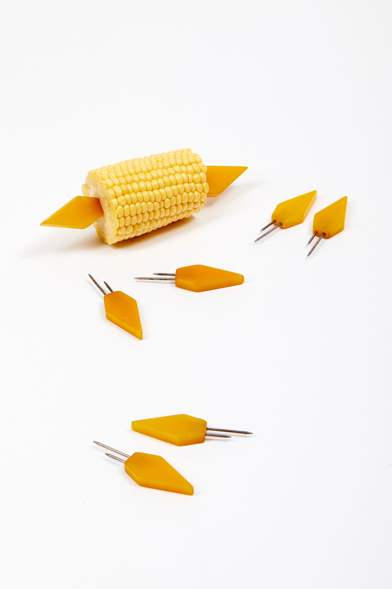 Set of yellow art-deco bakelite corn holders, photographed on a white background. One set is in a half a cob of corn. Three additional sets lay on the white surface. Corn holders are each a flat polygonal piece of yellow bakelite with two metal picks. 