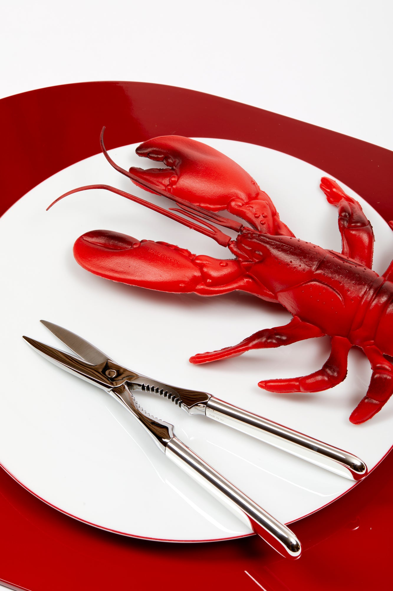 Silver lobster tongs resting aside a fake red lobster, both atop a white plate that is resting on a red lacquer tray.