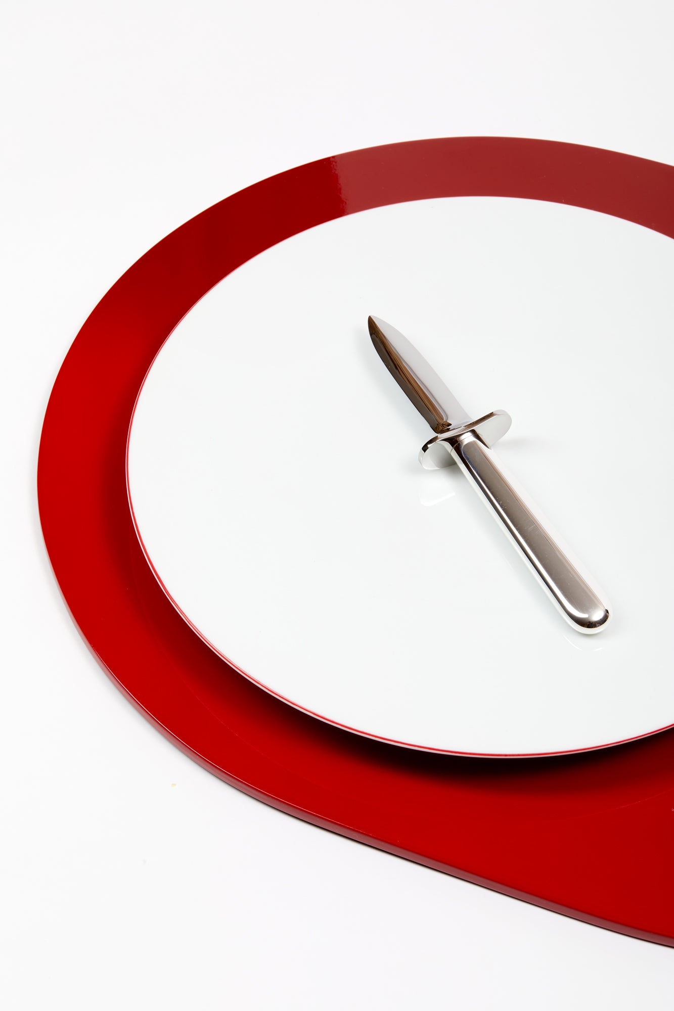 Silver oyster shucking knife resting atop a round white plate, atop a red serving tray.